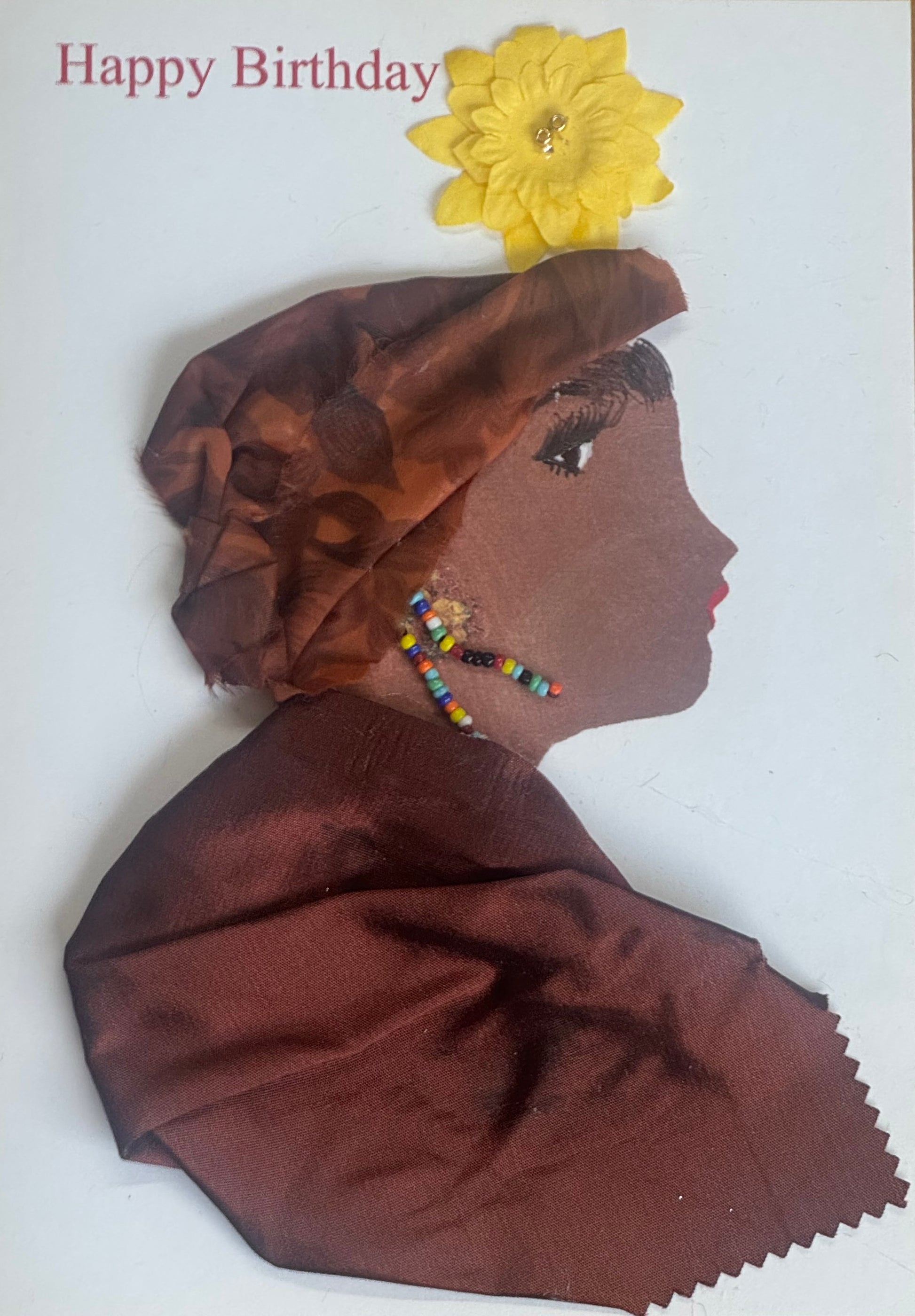 A handmade birthday card showing a side-profile illustration of a woman wearing a brown satin headwrap with a bright yellow flower embellishment at the top. She has long multicolored beaded earrings and a matching brown satin fabric garment forming her shoulders. Her lips are red and her eye is delicately drawn. The background is white with “Happy Birthday” printed above.