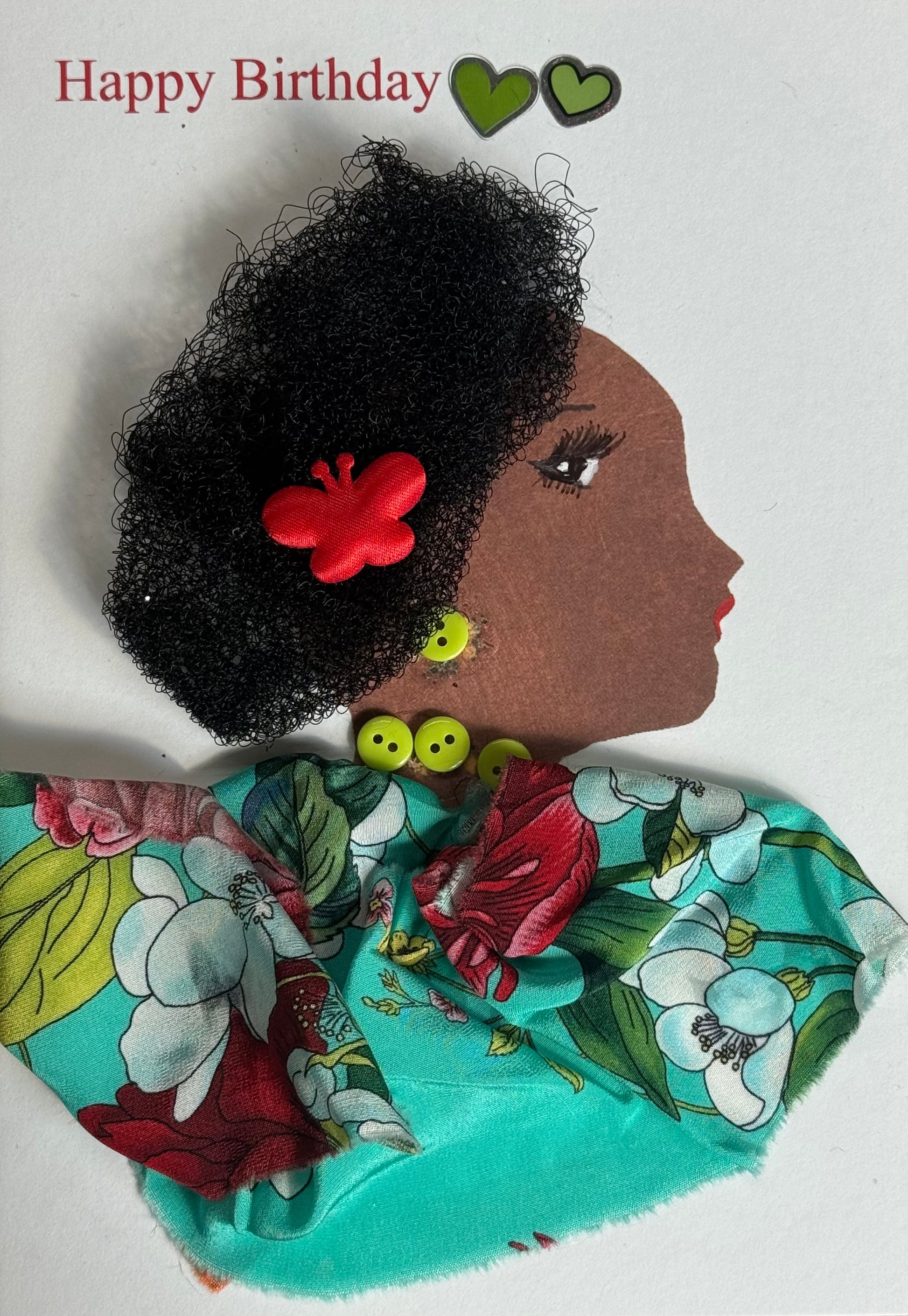 A handmade birthday card showing a woman with dark brown skin in right-facing profile. She has thick textured curly black hair with a red butterfly hair clip placed near the side. She wears lime green button earrings and green bead jewelry at her neck. Her clothing is made from bright tropical floral fabric in turquoise, red, green, and white tones draped across her shoulders. Two small green heart stickers appear near the top beside red “Happy Birthday” text on a white background.