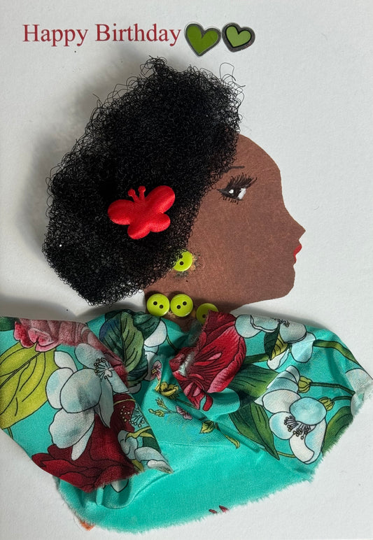 A handmade birthday card showing a woman with dark brown skin in right-facing profile. She has thick textured curly black hair with a red butterfly hair clip placed near the side. She wears lime green button earrings and green bead jewelry at her neck. Her clothing is made from bright tropical floral fabric in turquoise, red, green, and white tones draped across her shoulders. Two small green heart stickers appear near the top beside red “Happy Birthday” text on a white background.