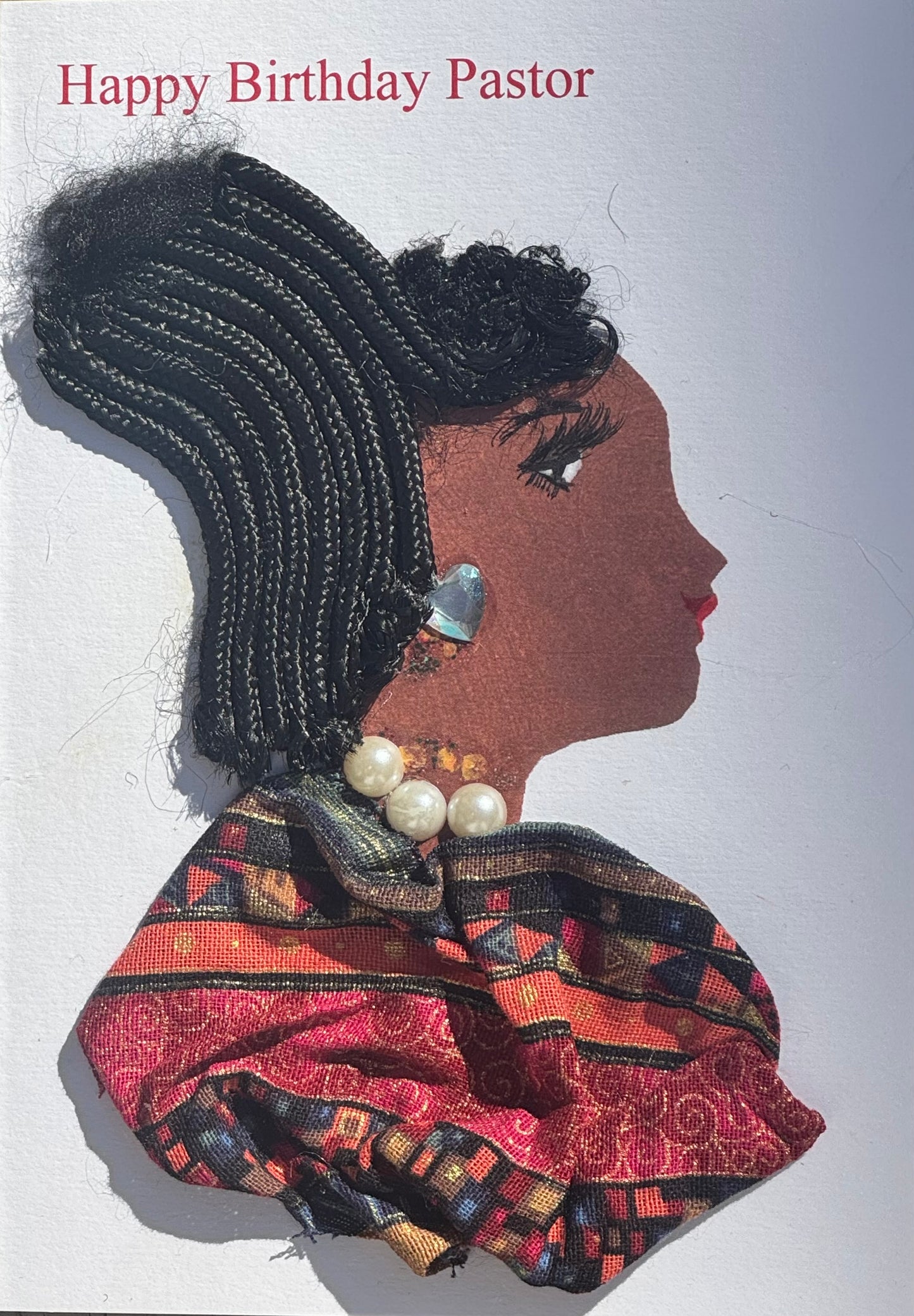 Handmade greeting card that can double as a piece of art featuring a womans  profile depicted with black braided hair detail worn in an updo. Shes also shown wearing pearl jewelry as a necklace, and a shinny silver earring in a shape of a heart. Her blouse is a patterned fabric garment, made up of red, orange, light blue, and black hues in different shapes. Above her head, theres also a “Happy Birthday Pastor” text toward the left side of the card. 
