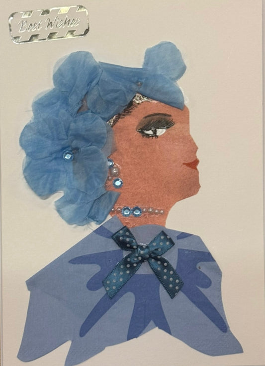 Handmade greeting card that doubles as a piece of art showing a woman in profile, with her hair formed from layered ligjt blue fabric flowers The floral layers vary in shade and texture. She wears a blue-toned outfit that blends seamlessly with the overall cool palette, with a patterned bow placed at the neckline as a central decorative detail. Pearl and gem accents are her matching neckalce and earrings set. Also above her head to the upper right is a sticker that reads "Best Wishes" in shiny silver. 
