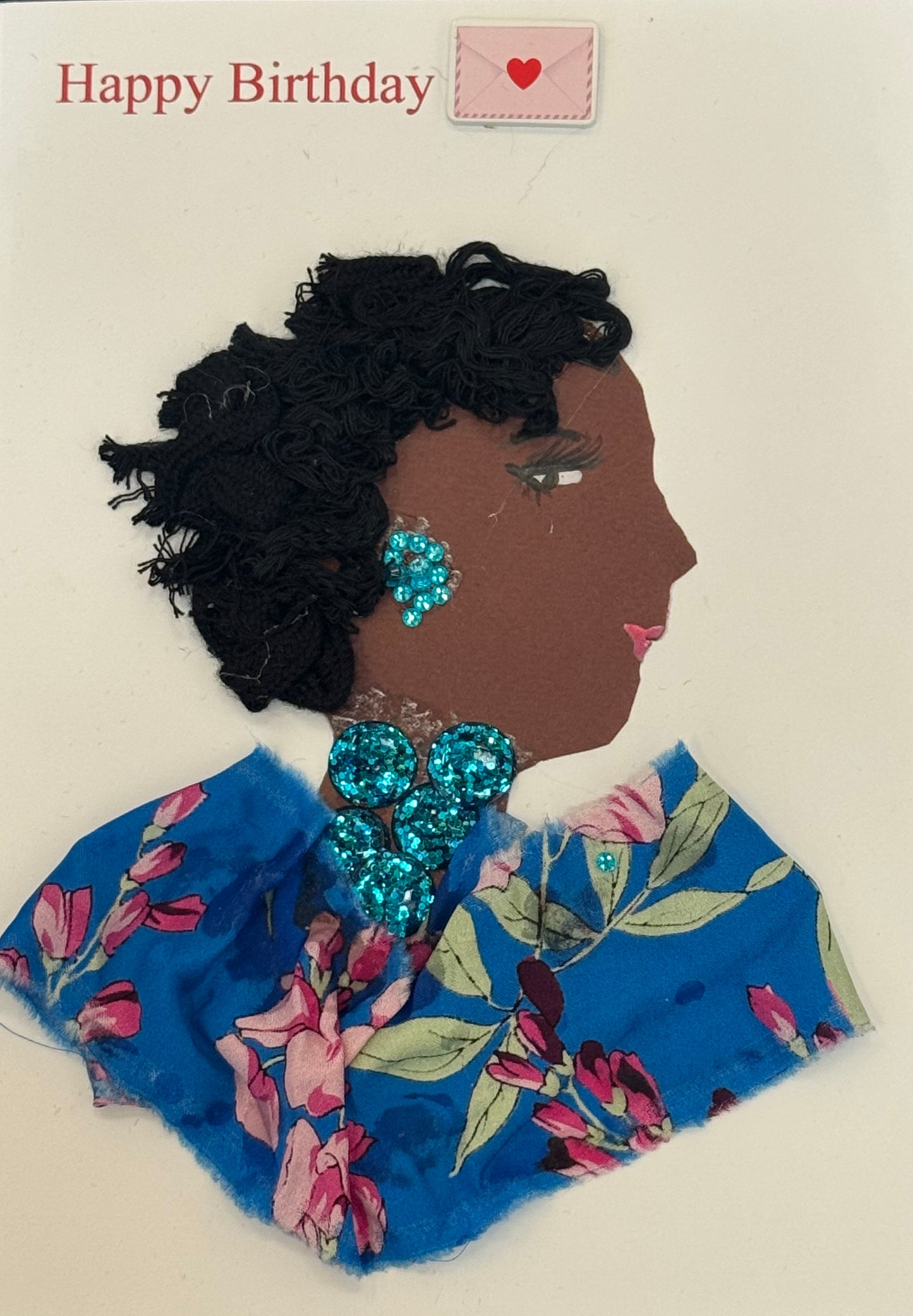 Handmade collage greeting card depicting a right-facing side profile of a dark-skinned woman with voluminous, textured black hair styled outward in a natural shape. Her face includes defined eyelashes, a shaped eyebrow, and soft pink lips. She wears large, bright turquoise-blue beaded earrings and a matching clustered statement necklace made of round, reflective beads. Her clothing is made from blue fabric featuring a floral print with pink flowers and green leaves. It also says “Happy Birthday" above. 