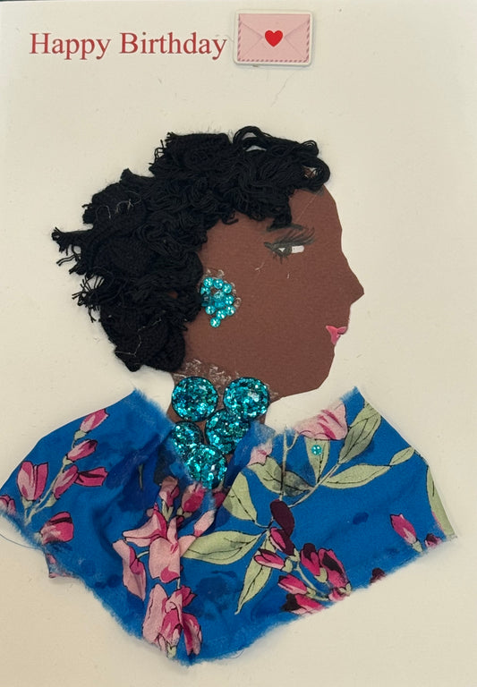 Handmade collage greeting card depicting a right-facing side profile of a dark-skinned woman with voluminous, textured black hair styled outward in a natural shape. Her face includes defined eyelashes, a shaped eyebrow, and soft pink lips. She wears large, bright turquoise-blue beaded earrings and a matching clustered statement necklace made of round, reflective beads. Her clothing is made from blue fabric featuring a floral print with pink flowers and green leaves. It also says “Happy Birthday" above. 