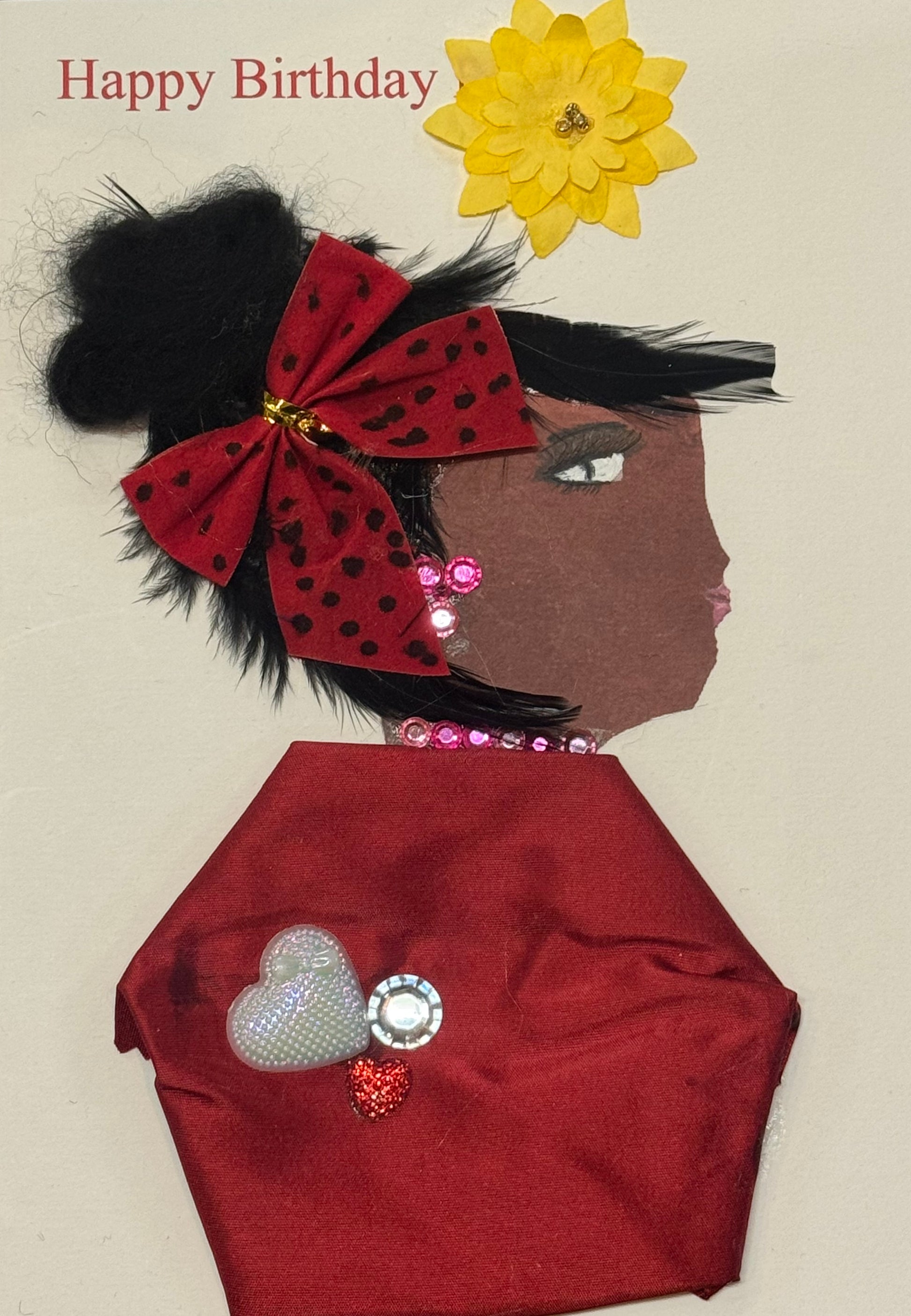 Handmade greeting card that can double as a piece of art featuring a side-profile portrait of a woman with a bold red bow with black polka dots in her dark hair, wearing a deep red satin-textured outfit adorned with sparkling heart embellishments. The card includes a bright yellow flower and “Happy Birthday” text at the top, combining fabric, rhinestones, and paper elements to create a vibrant, celebratory, and textured design.