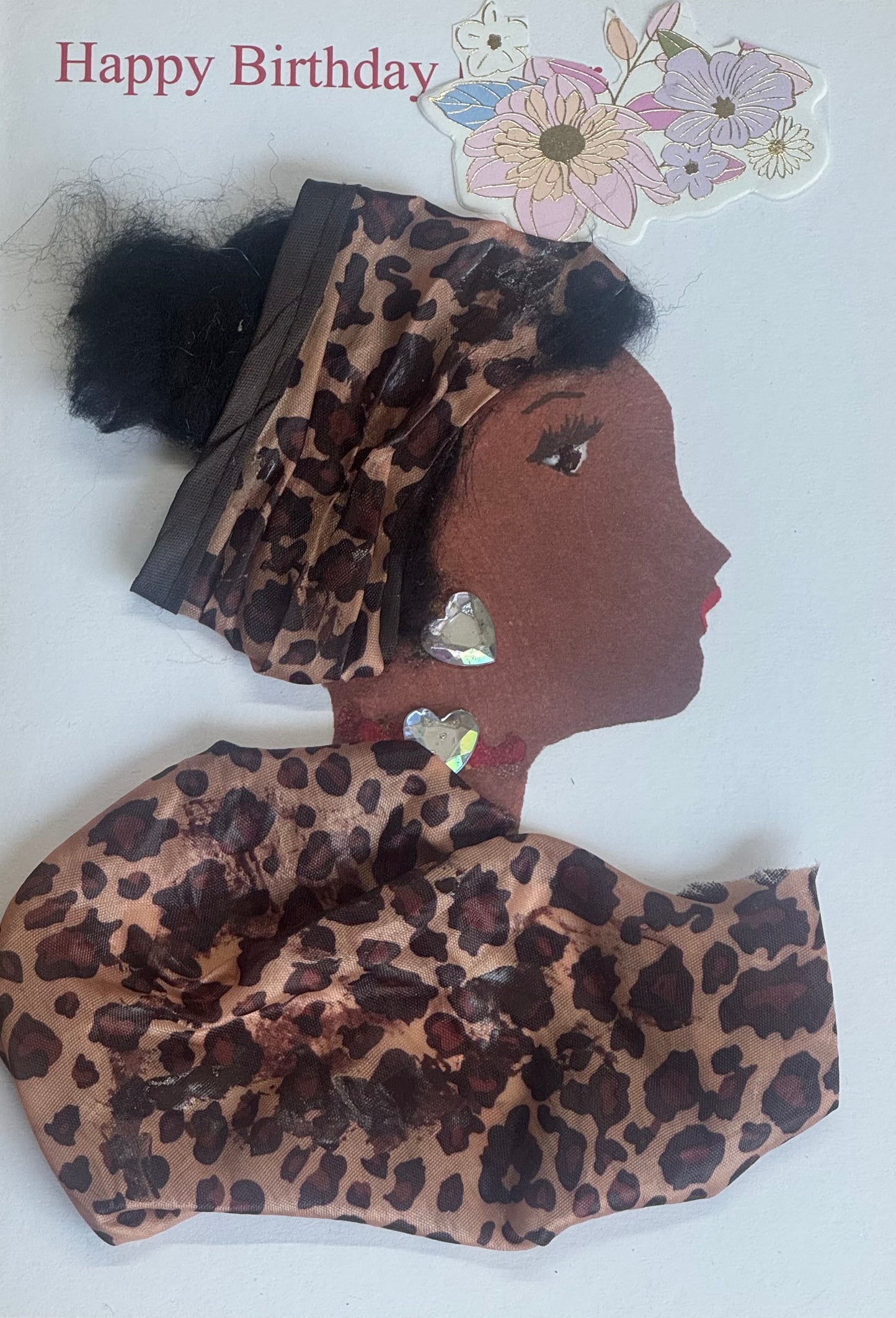 A handmade birthday card showing a woman in profile wearing a leopard print fabric headwrap and matching garment. She has large heart-shaped gem earrings and a matching heart gem necklace. Her hair is styled into two puff buns behind the wrap. The background is white with “Happy Birthday” text and floral stickers in the top corner.