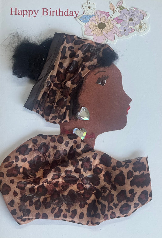 A handmade birthday card showing a woman in profile wearing a leopard print fabric headwrap and matching garment. She has large heart-shaped gem earrings and a matching heart gem necklace. Her hair is styled into two puff buns behind the wrap. The background is white with “Happy Birthday” text and floral stickers in the top corner.