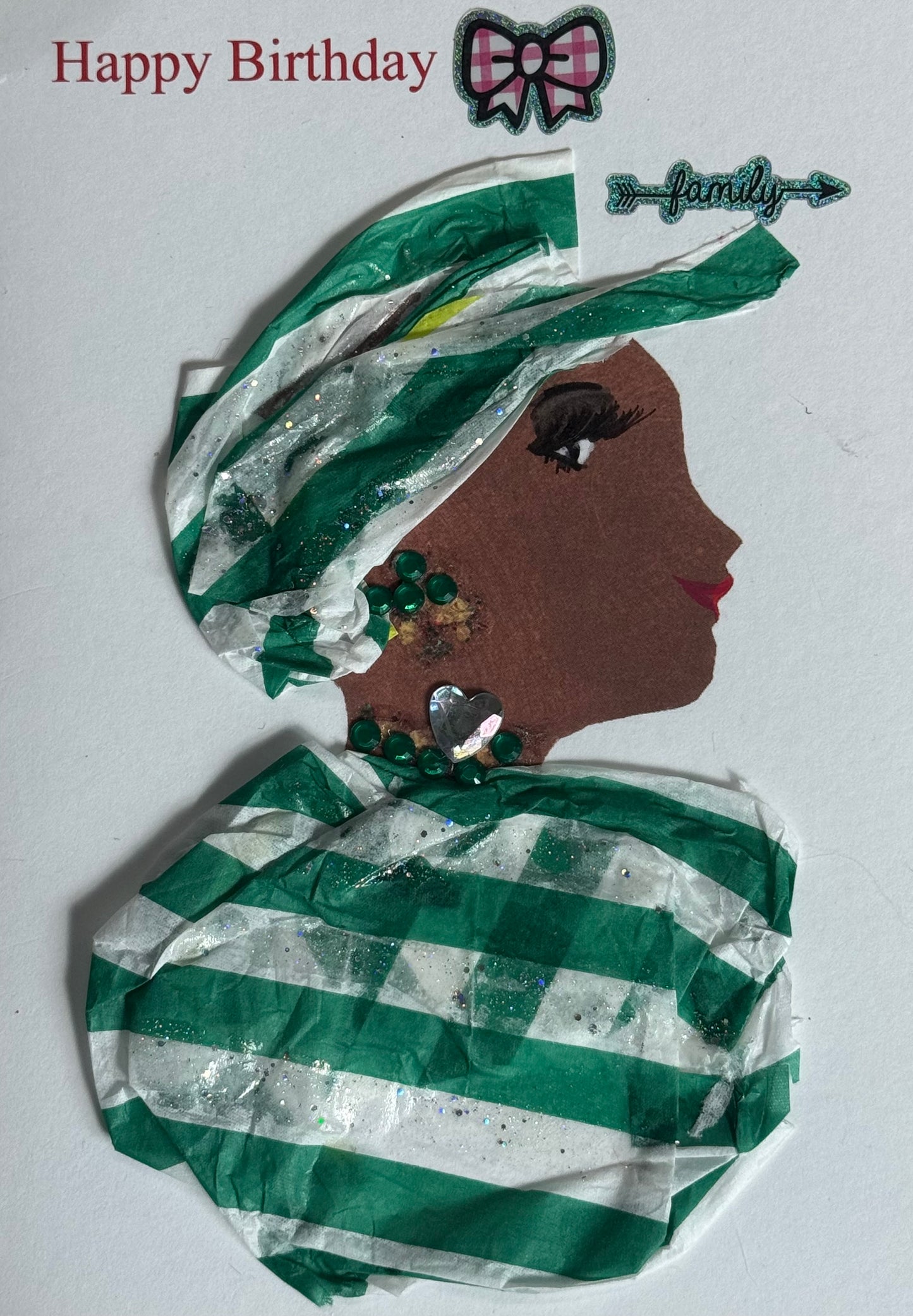 A handmade birthday card featuring a woman with dark brown skin in side profile facing right. She wears a layered green and white striped headwrap with glitter accents. Her jewelry includes green gem earrings and a silver heart-shaped pendant necklace. Her shoulders are wrapped in matching green and white striped fabric. Above her head are a bow sticker and decorative “family” accent, with “Happy Birthday” printed in red on a white background