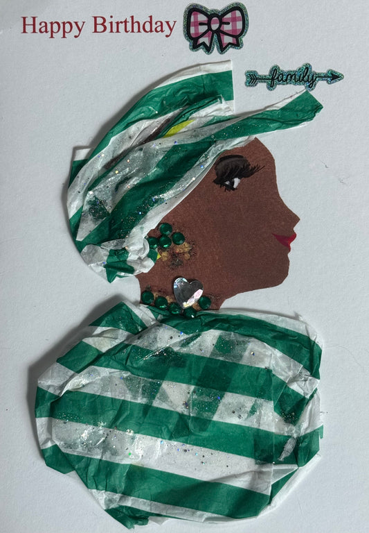 A handmade birthday card featuring a woman with dark brown skin in side profile facing right. She wears a layered green and white striped headwrap with glitter accents. Her jewelry includes green gem earrings and a silver heart-shaped pendant necklace. Her shoulders are wrapped in matching green and white striped fabric. Above her head are a bow sticker and decorative “family” accent, with “Happy Birthday” printed in red on a white background