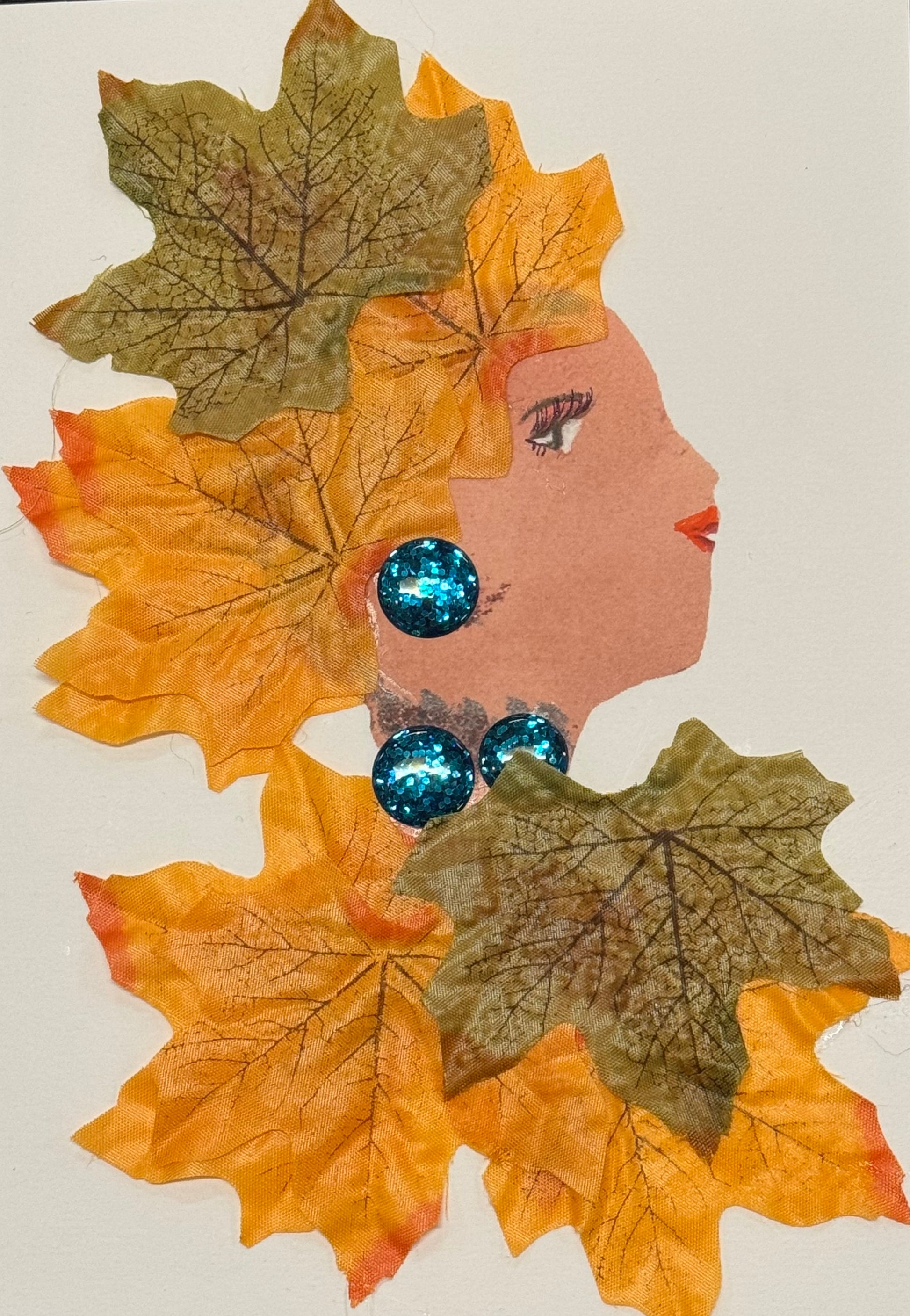 Handmade greeting card that doubles as a piece of art featuring a side-profile collage of a woman composed from layered autumn leaves. The leaves, in shades of orange, golden yellow, and deep brown, are arranged to form both her flowing hair and parts of her clothing, creating a textured, organic silhouette. The overlapping leaf shapes give the card a sense of depth, as if the hair is gently cascading around her shoulders. She wears large, bright blue gem earrings that stand out against the warm tones. 