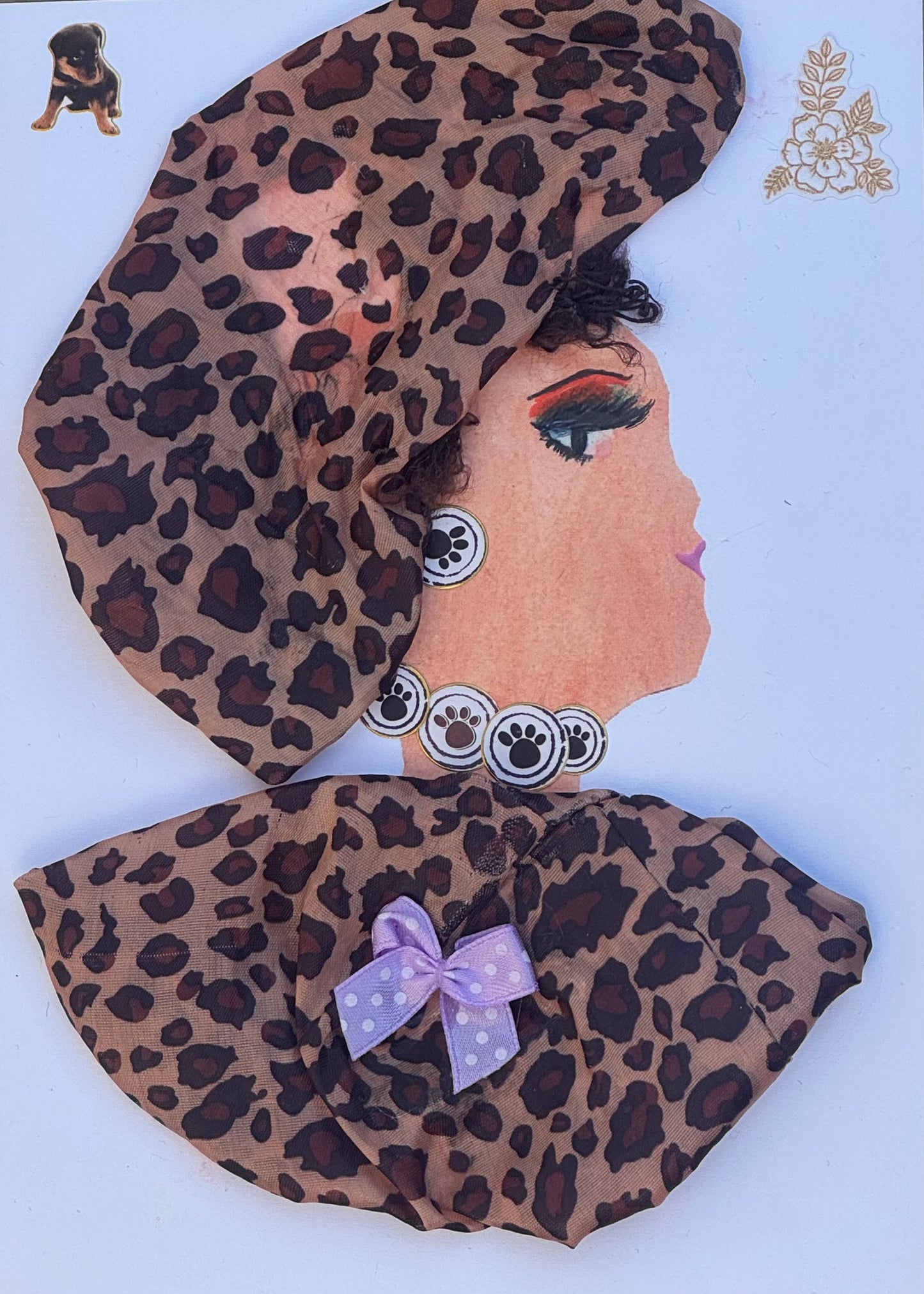 Handmade greeting card that can double as a piece of art featuring a woman in profile wearing a large cheetah print headwrap with soft fabric folds and a matching patterned garment. She has bold eye makeup, a stylized painted eye, and a decorative necklace made from circular elements resembling paw-print motifs. Small cat stickers appear near the top corner, and a lavender bow embellishment is attached to the garment, adding a playful and layered mixed-media effect with multiple textures.