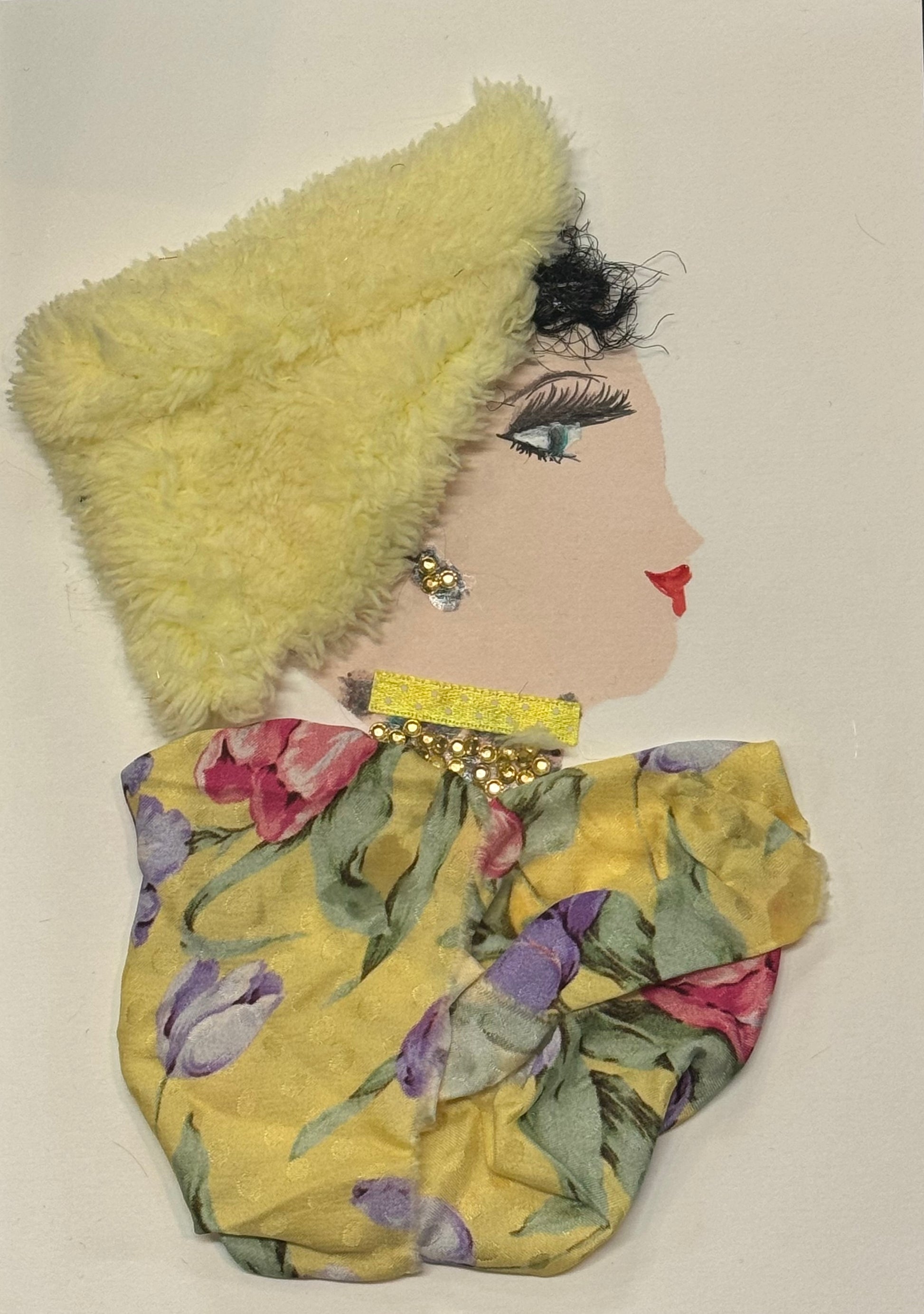 Handmade greeting card that can double as a piece of art featuring a side-profile portrait of a woman wearing a soft, fuzzy pale-yellow hat made from textured fabric and a floral-patterned outfit in muted colors. The design includes small gold embellishments at the neckline, delicate facial details with long lashes and red lips, and a combination of soft textiles and paper elements that create a cozy and elegant mixed-media look.