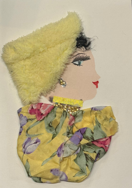 Handmade greeting card that can double as a piece of art featuring a side-profile portrait of a woman wearing a soft, fuzzy pale-yellow hat made from textured fabric and a floral-patterned outfit in muted colors. The design includes small gold embellishments at the neckline, delicate facial details with long lashes and red lips, and a combination of soft textiles and paper elements that create a cozy and elegant mixed-media look.