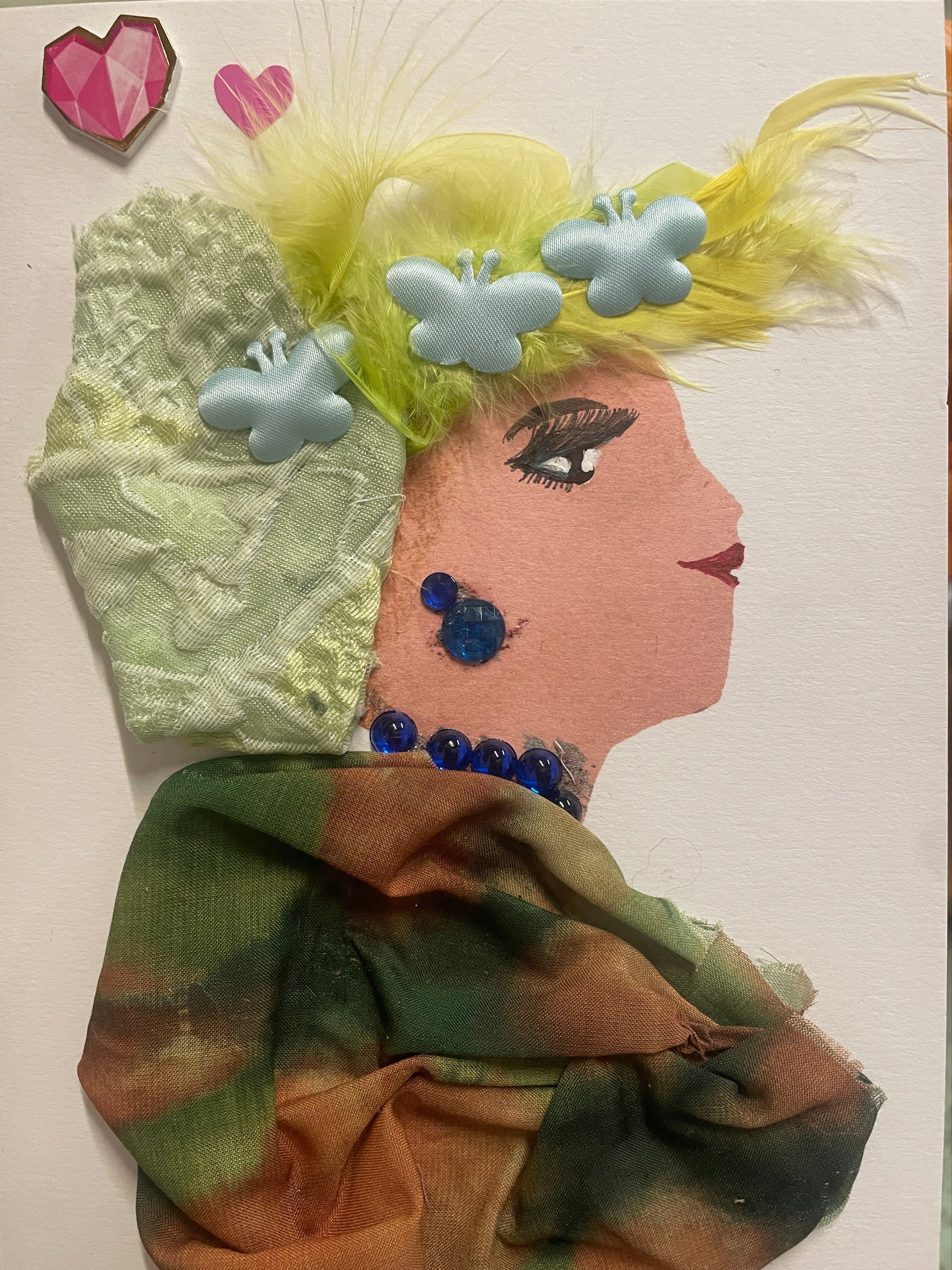 A handmade card shows a side-profile portrait of a woman with pale skin, red lips, and dramatic black lashes. She has fluffy yellow hair decorated with pastel blue butterfly embellishments and feathers. She wears large blue gem earrings, a matching blue beaded necklace, and a textured green-and-brown fabric wrap. A pink heart and a pink geometric heart sticker appear in the corner.