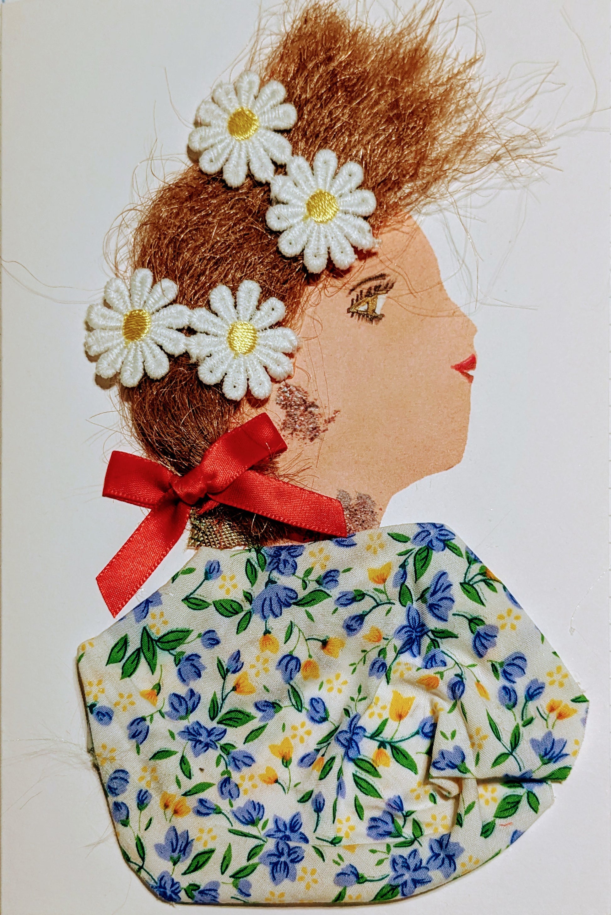 This is a handmade card of a woman named Chamomile Daisy. Her hair is styled with soft brown fibres and decorated with five white daisies. A red ribbon is tied at the back of her neck. She wears a fabric top covered in a delicate floral pattern with blue, yellow, and green blossoms on a light background. 