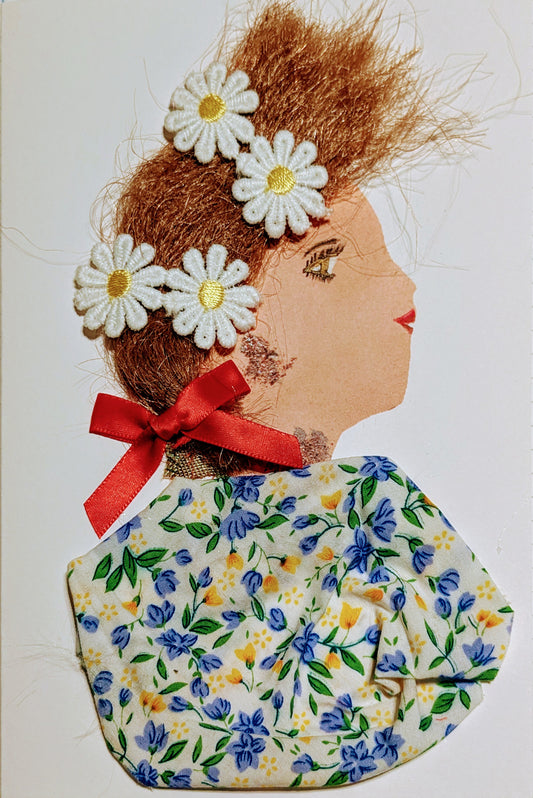 This is a handmade card of a woman named Chamomile Daisy. Her hair is styled with soft brown fibres and decorated with five white daisies. A red ribbon is tied at the back of her neck. She wears a fabric top covered in a delicate floral pattern with blue, yellow, and green blossoms on a light background. 