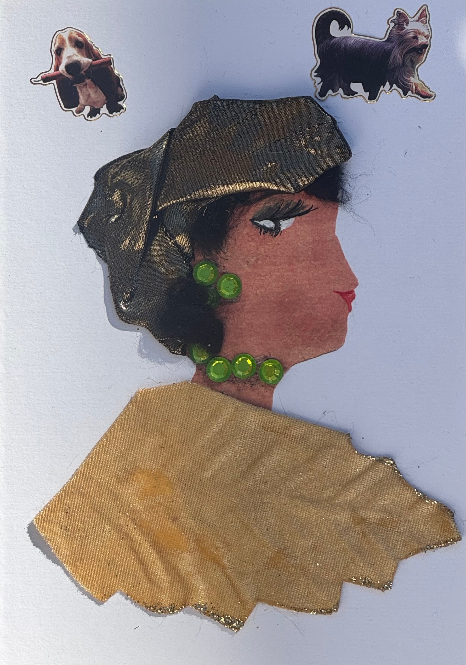 Handmade greeting card that can double as a piece of art featuring a woman in profile wearing a dark gold shimmery headwrap. She is also pictured wearing green jewel earrings with a matching necklace. For her blouse, it is shapped in the figure of a leaf that is made up of light velvet texture and is a light beige/gold colour, with the outline of the blouse lined in gold glitter. Also above on each corner of the card are dog stickers placed. 