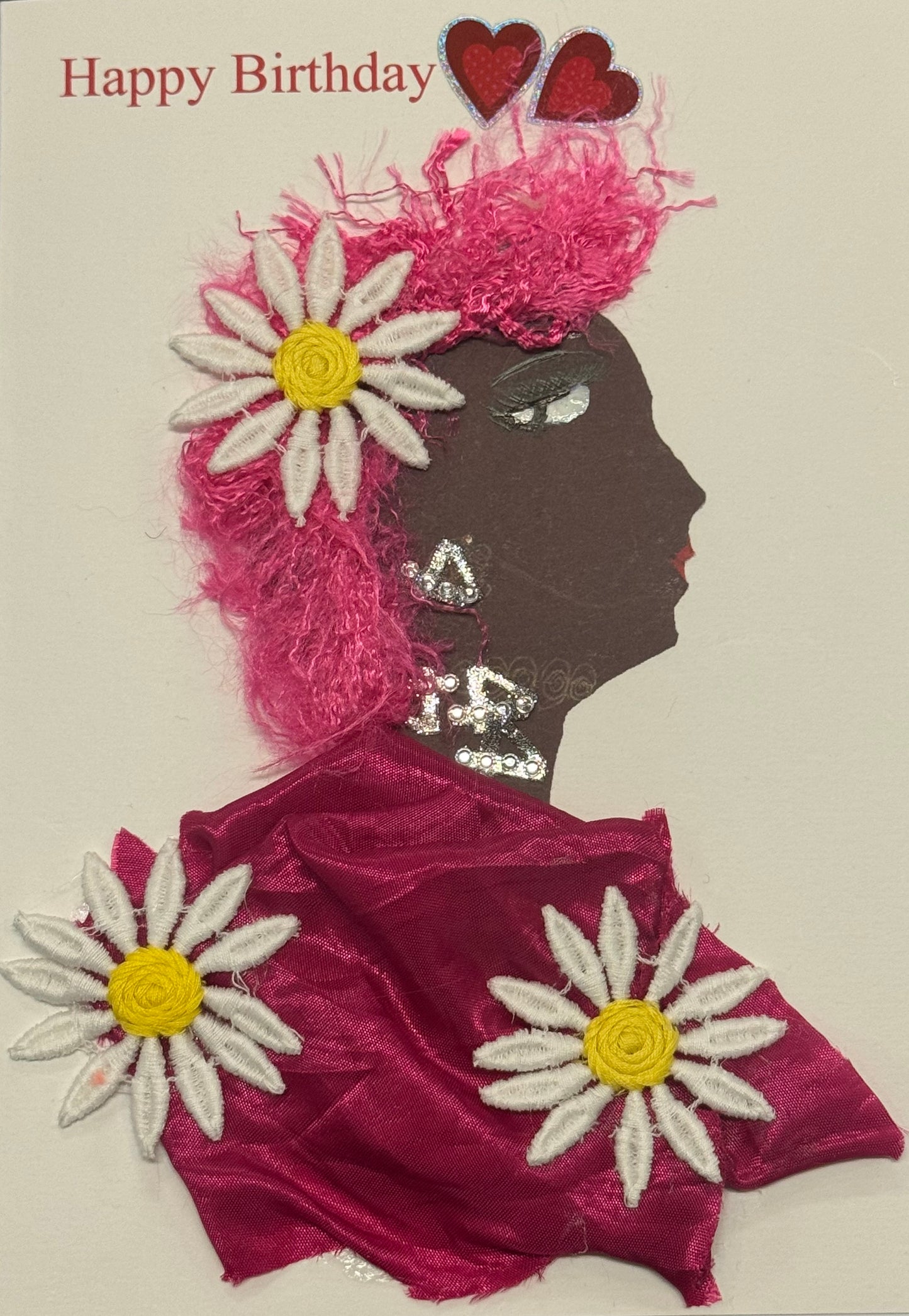 Handmade greeting card that can double as a piece of art, featuring a side-profile collage of a woman with brown skin facing left, featuring bold pink textures. Her hair or head covering appears full and textured in bright pink tones. Large white daisy flowers with yellow centers are placed prominently around her head and shoulder area. Above her, small pink heart graphics float, adding a playful and cheerful feel. The image is bright, colorful, and full of layered decorative elements