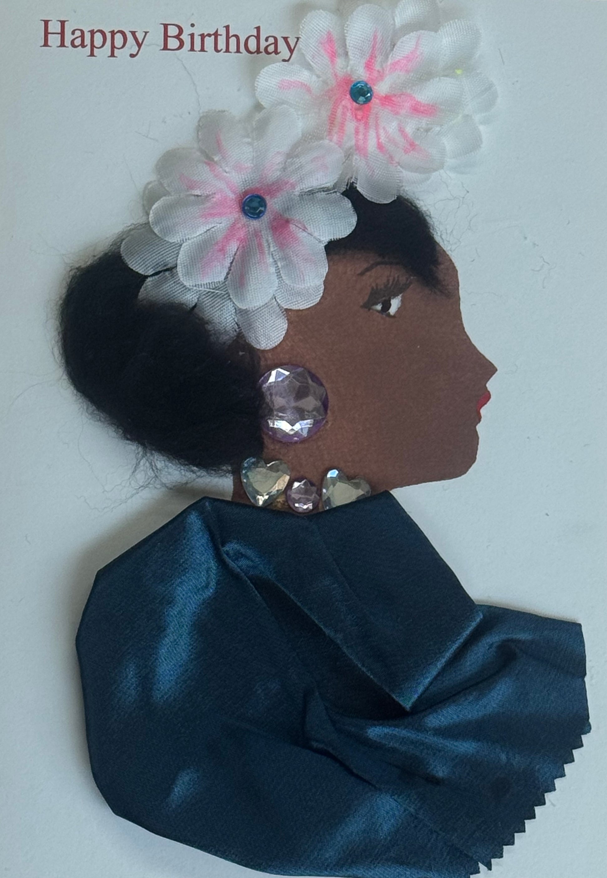 A handmade birthday card showing a side-profile illustration of a woman wearing two white flower embellishments in her hair. She has large purple gem earrings and smaller heart gems along her neckline. Her clothing is made from teal satin fabric. The background is white with “Happy Birthday” text above.