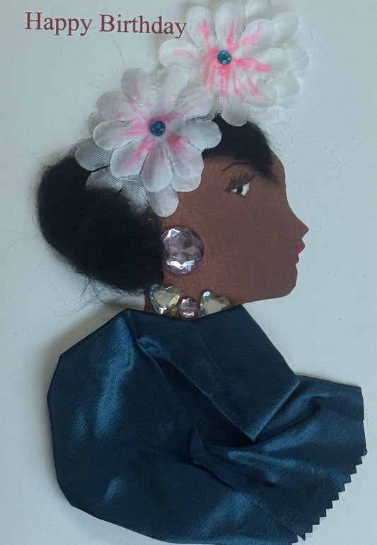 A handmade birthday card showing a side-profile illustration of a woman wearing two white flower embellishments in her hair. She has large purple gem earrings and smaller heart gems along her neckline. Her clothing is made from teal satin fabric. The background is white with “Happy Birthday” text above.
