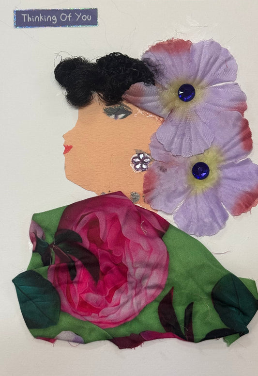 Handmade greeting card that can double as a piece of art, featuring a woman with dark textured hair adorned with large purple flowers. She wears a floral-patterned garment with a prominent rose design and gem accents, with a “Thinking of You” sticker in the top right corner above her head. 