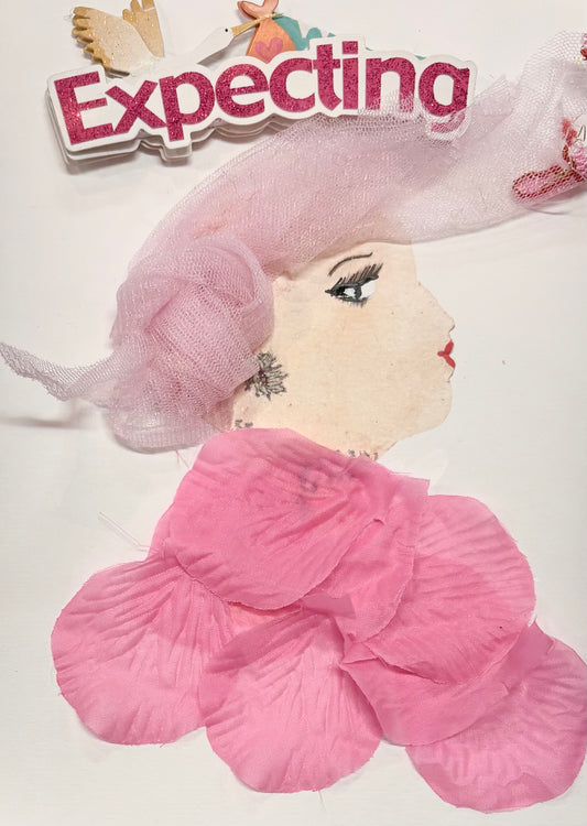 A woman wears a light pink hat made of tulle. Her blouse is a slightly darker pink made of petals. She wears subtle white earring. At the top of the card, a sticker says "Expecting" with a stork flying above. 