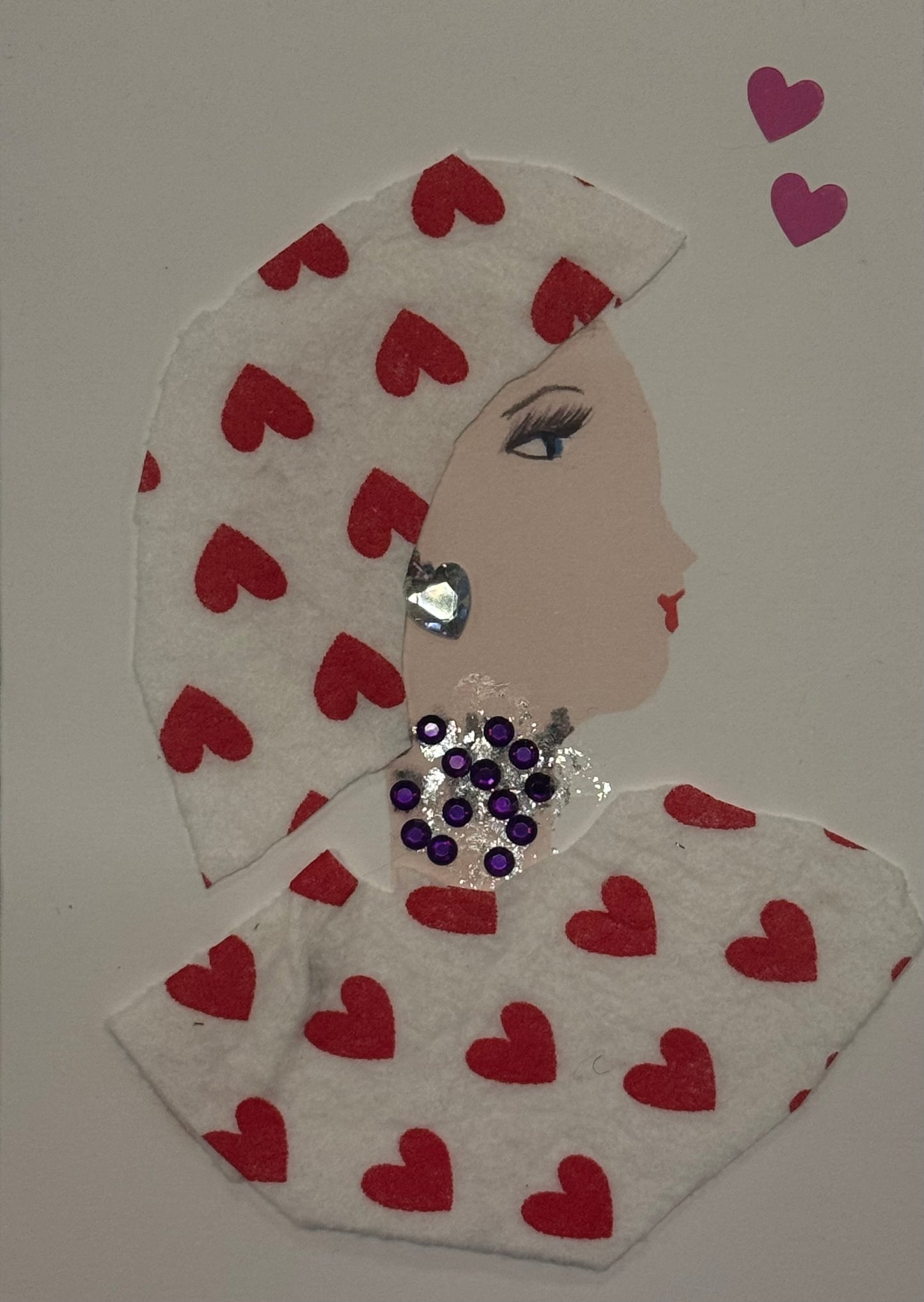 A handmade card that can double as an art piece showing a profile of a woman wearing a white felt headwrap and shawl, both patterned with red hearts. She is decorated with a purple rhinestone necklace and a silver heart-shaped earring.