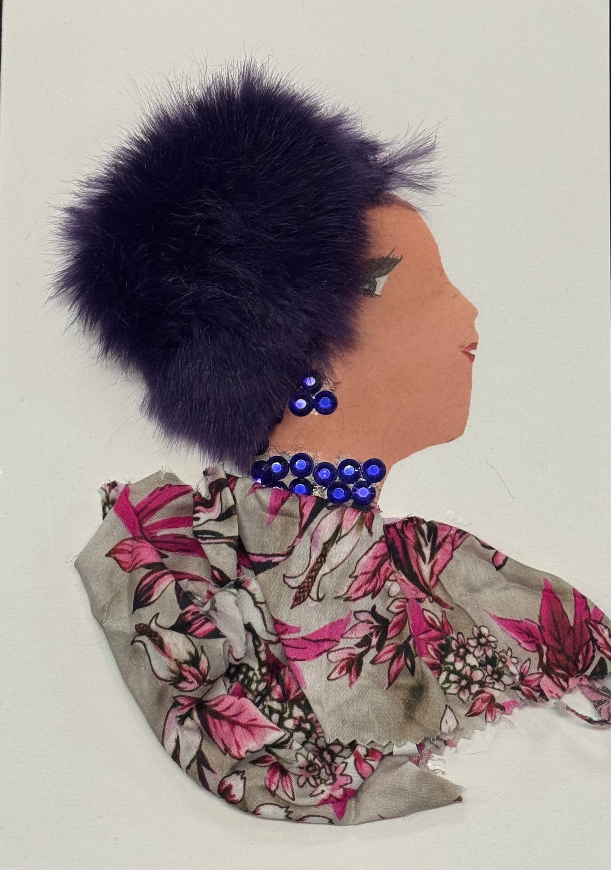 Handmade greeting card that can double as a piece of art, a side-profile mixed media collage of a woman with a medium skin tone set against a white background. She wears a voluminous purple faux-fur hat with a soft, fluffy texture that frames her head. Her outfit is made of a floral-patterned fabric featuring pink flowers and neutral tones, gathered around the neckline. Bright blue circular beads form a necklace and matching earring. Her face is illustrated with a defined eye, long lashes, and a subtle lip.