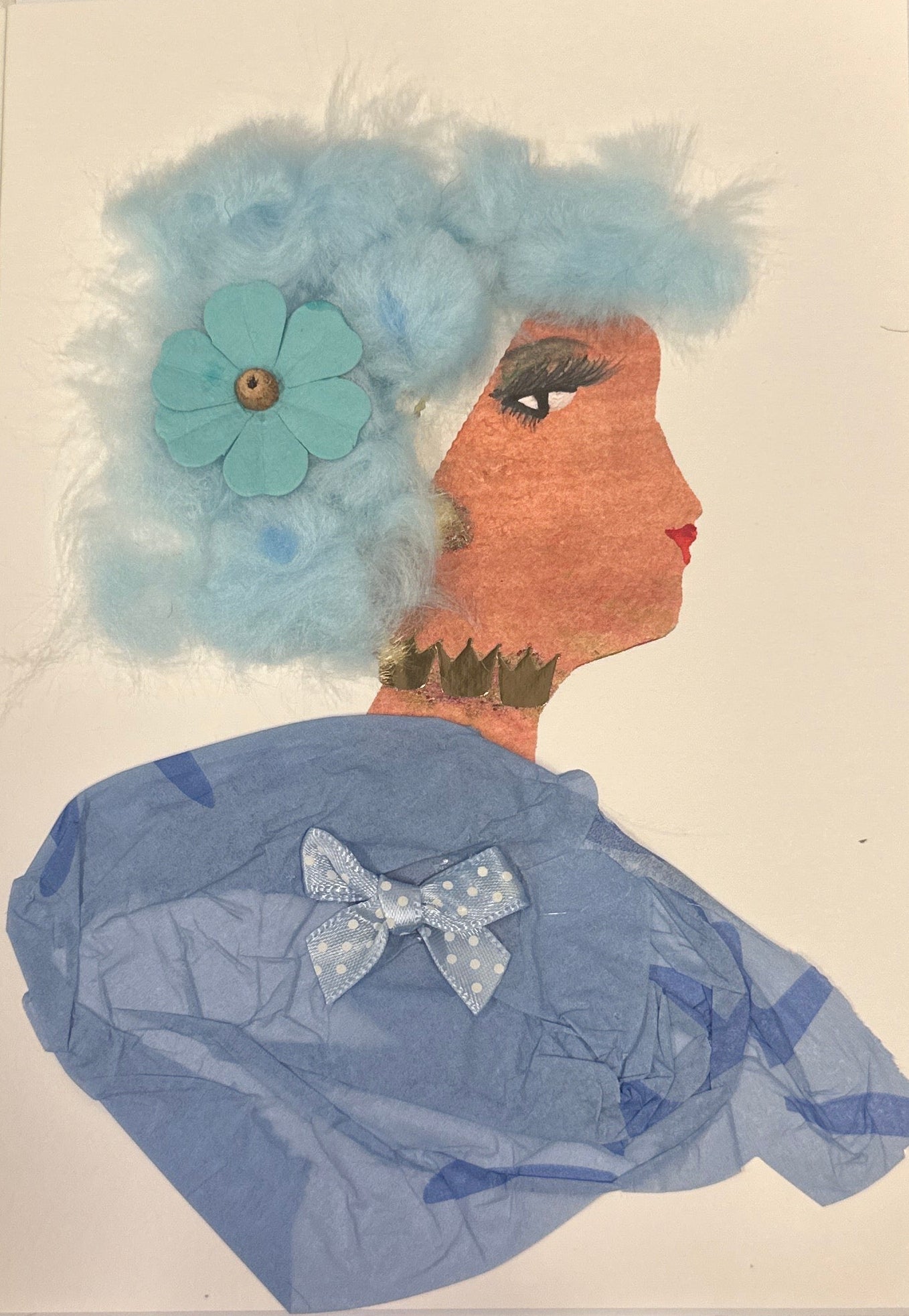 A handmade collage of a woman in profile with light blue fluffy cotton hair decorated with a blue flower. She has warm brown skin, long dark eyelashes, and bright red lipstick. She wears gold jewelry around her neck and a light blue textured blouse made from tissue paper with a polka-dot bow on the front. The background is plain white.