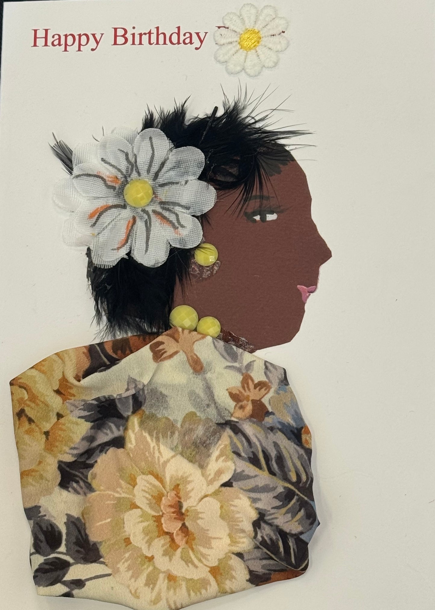 Handmade collage greeting card featuring a right-facing side profile of a woman with medium to dark brown skin. She has short black hair styled with a soft, feathery texture. A large white fabric flower with a yellow center is placed near her temple, accompanied by black feather accents. Her facial features include a defined eye with lashes and soft pink lips. She wears round yellow bead earrings and a coordinating necklace. The background is white with “Happy Birthday” printed in red text and a small daisy