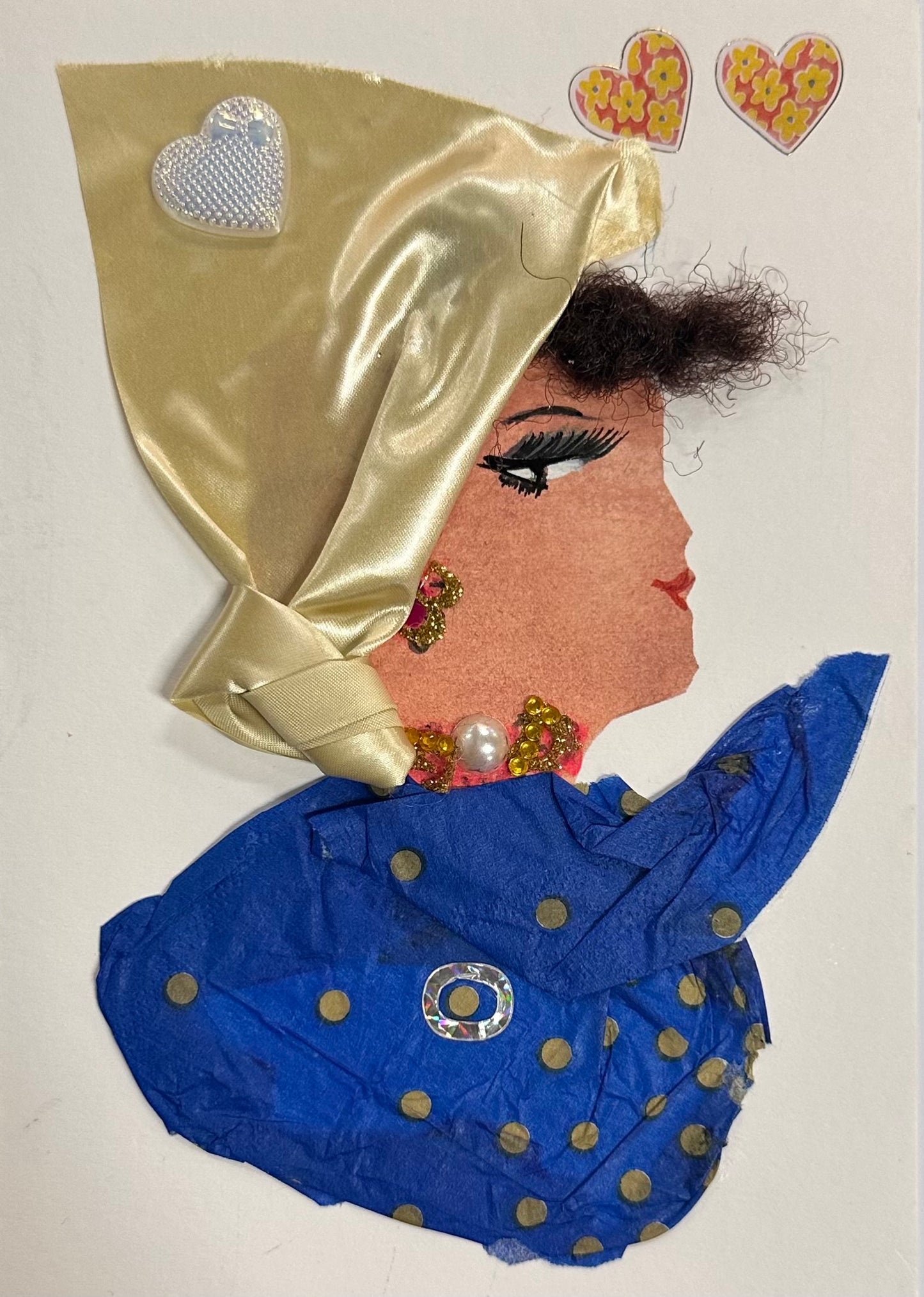 A handmade collage portrait of a woman in profile featuring a gold satin headscarf, curly dark hair, dramatic eyelashes, and red lipstick. She wears a royal-blue polka-dot outfit and pearl-and-gem jewellery, with decorative heart stickers above her head.