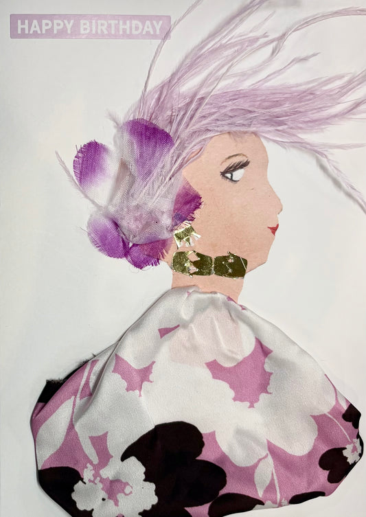 This is a handmade card of a woman named **Fuchsia Feather**. Her flowing lavender feathers sweep to the side, accented with delicate purple and white blossoms. She wears a voluminous dress in a silky white fabric patterned with large floral prints in soft pink, deep plum, and white. Her look is finished with bold, textured gold accessories, and a “Happy Birthday” message appears in the top corner in soft purple.