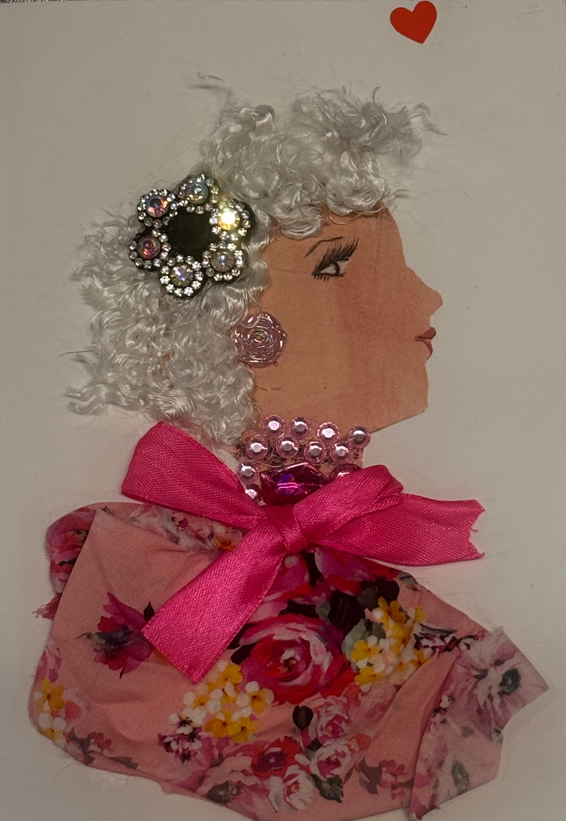 Princess Silver Street is depicted here with white curly textured hair. She has a gillmering silver clip in hair as well as her shiny pink jewellry. She is wearing rose shaped earrings and a complimentary pink beaded necklace. On her light pink blouse, she has a bright floral print that features flowers twith hues of red, pink, yellow, dark green, and white. There is also a small detailed red heart on the top right of the card that adds a nice final touch.