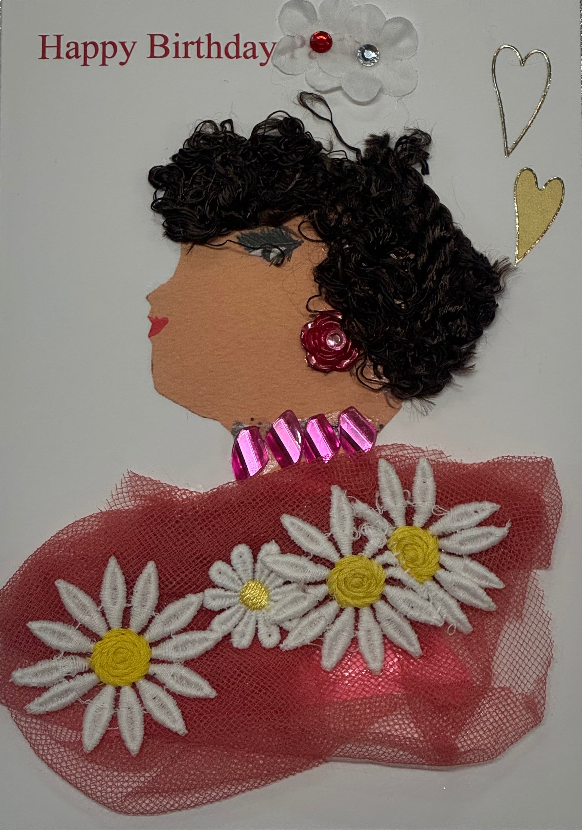 Handmade birthday card that can double as a piece of art showing a woman with brown curly hair. She wears a warm pink tulle textured blousewith four white and yellow daisy flowers embroideredonto the shirt. She has pink colored jewelry and decorative gems with red rose shaped earrings, creating a cheerful and colorful celebration theme. She is also surrounded by two white flowers and two white and gold hearts above her head to the top right of the card. "Happy Birthday" is also written across the top left. 