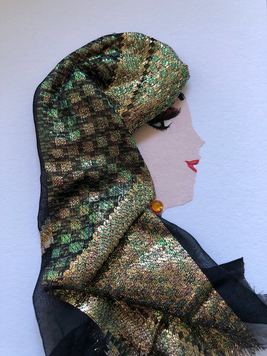This is a handmade card of a woman named Gold Guatemala. She wears a shimmering gold and green headscarf that wraps around her head and shoulders, made from textured metallic fabric with hints of black. A small amber gem sits just below her ear like an earring. Her lips are painted red in a soft smile, and her eye is accentuated with long, dramatic lashes. The overall look is elegant and regal.