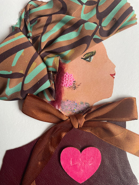 This is a handmade card of a woman named Holland Park Holly. She wears a bright head wrap with brown, teal, and green stripes. Her earring is a small pink flower, and her neck sparkles with glitter. She’s dressed in burgundy fabric with a shiny brown ribbon tied in a bow. A bold pink heart on her chest reads “Love.”