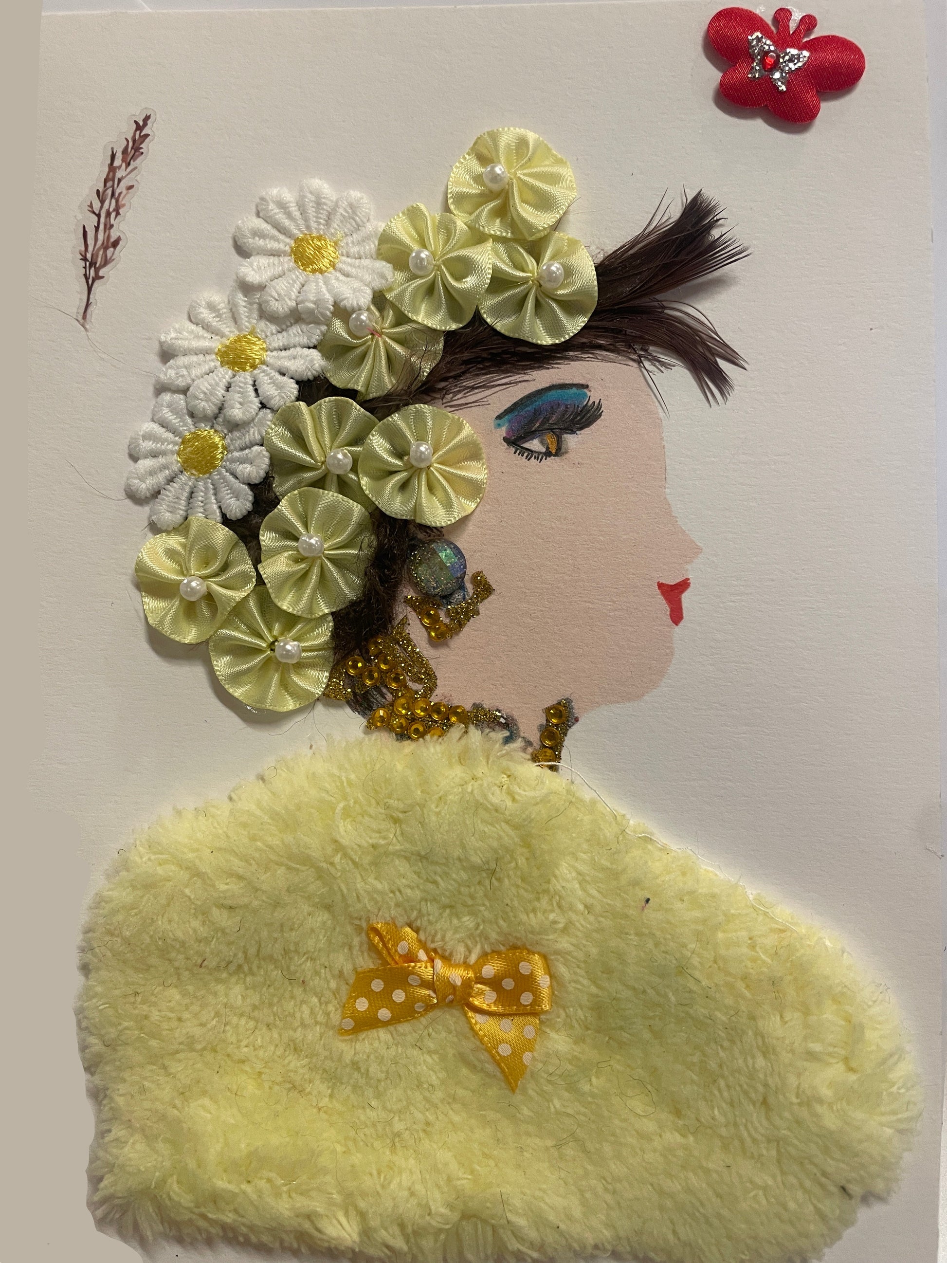 A handmade card features a side-profile portrait of a woman with light skin, red lips, and dramatic blue-and-yellow eye makeup. Her hair is decorated with a crown of fabric flowers in white and pale yellow, each with pearl or yellow button centers. She wears beaded gold jewelry and a large fuzzy yellow shawl with a small polka-dotted yellow bow attached. A red butterfly embellishment and a sprig-like decoration appear near the top of the card.