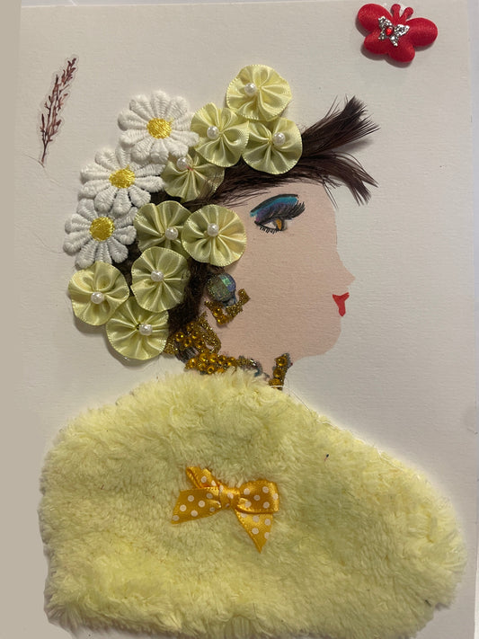 A handmade card features a side-profile portrait of a woman with light skin, red lips, and dramatic blue-and-yellow eye makeup. Her hair is decorated with a crown of fabric flowers in white and pale yellow, each with pearl or yellow button centers. She wears beaded gold jewelry and a large fuzzy yellow shawl with a small polka-dotted yellow bow attached. A red butterfly embellishment and a sprig-like decoration appear near the top of the card.