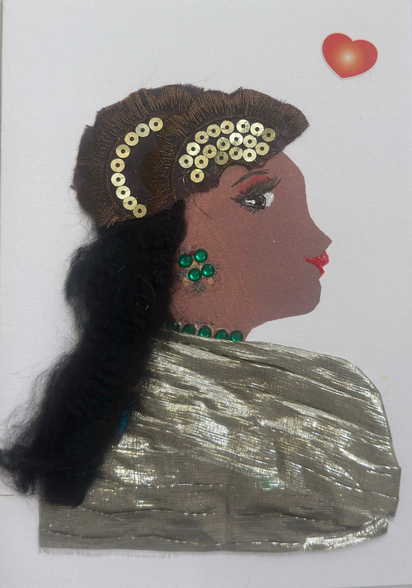 A handmade card shows a side-profile portrait of a woman with deep brown skin, long black textured hair, and red lips. She wears golden sequins in her hair, green jeweled earrings, and a green necklace. Her outfit is made of shimmering silver fabric, and a small golden heart sticker appears near the top.