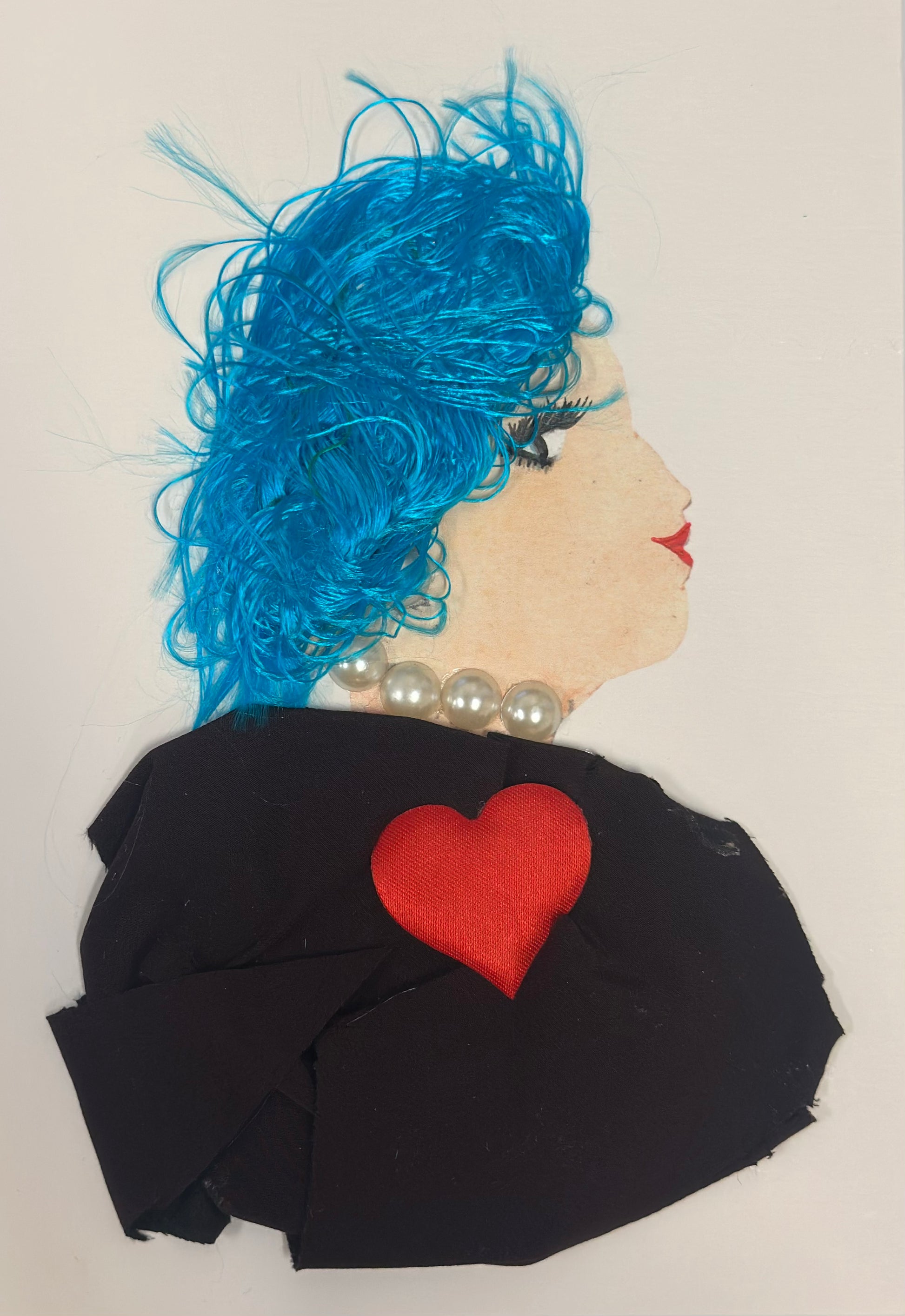 A handmade card shows a side-profile portrait of a woman with pale skin, bright electric-blue textured hair, and red lips. She wears a pearl necklace and a black fabric outfit adorned with a red heart shape on the chest. The overall look is bold, stylish, and energetic.