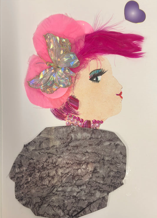 A handmade card shows a side-profile portrait of a woman with pale skin, pink feathered hair, and teal eye makeup. A large iridescent butterfly rests on a pink fabric flower in her hair, and she wears magenta gem earrings and a matching pink bead necklace. A grey, textured fabric drapes across her shoulders, and a purple heart sticker sits near the top corner.
