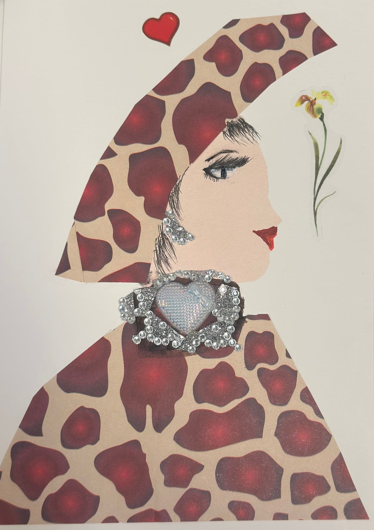 A handmade card shows a side-profile portrait of a woman with light skin, black eyelashes, and red lips. She wears a large giraffe-print hat and matching outfit, along with silver jeweled earrings and a prominent silver heart brooch made of rhinestones. A small red heart and a delicate flower sticker appear near her head on the card.