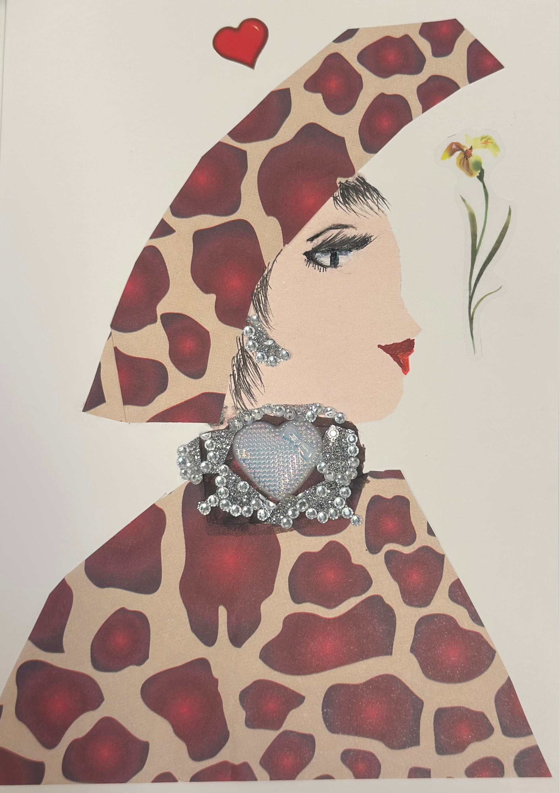 A handmade card shows a side-profile portrait of a woman with light skin, black eyelashes, and red lips. She wears a large giraffe-print hat and matching outfit, along with silver jeweled earrings and a prominent silver heart brooch made of rhinestones. A small red heart and a delicate flower sticker appear near her head on the card.