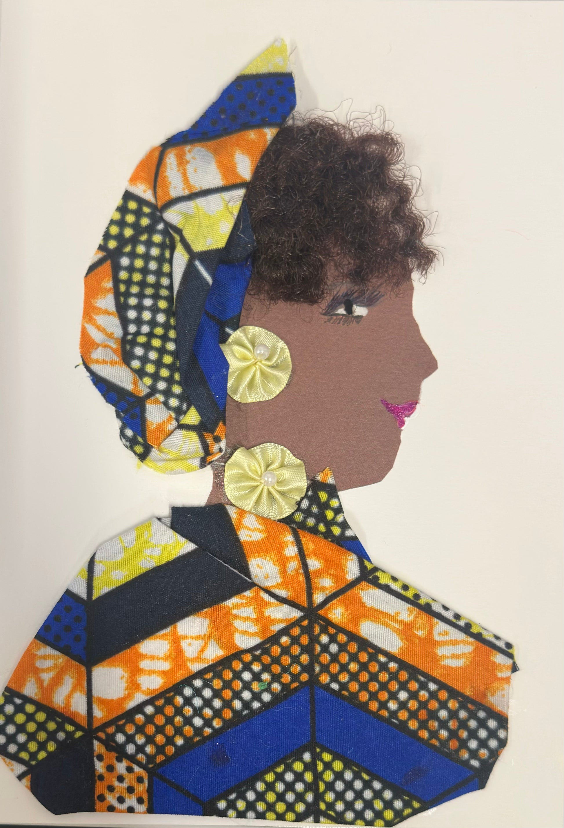 A handmade card features a side-profile portrait of a woman with deep brown skin, curly brown textured hair, and pink lips. She wears a colourful geometric-patterned headwrap and matching outfit in blue, orange, black, and yellow tones. Decorative pale-yellow satin flower embellishments serve as earrings and a collar accent.