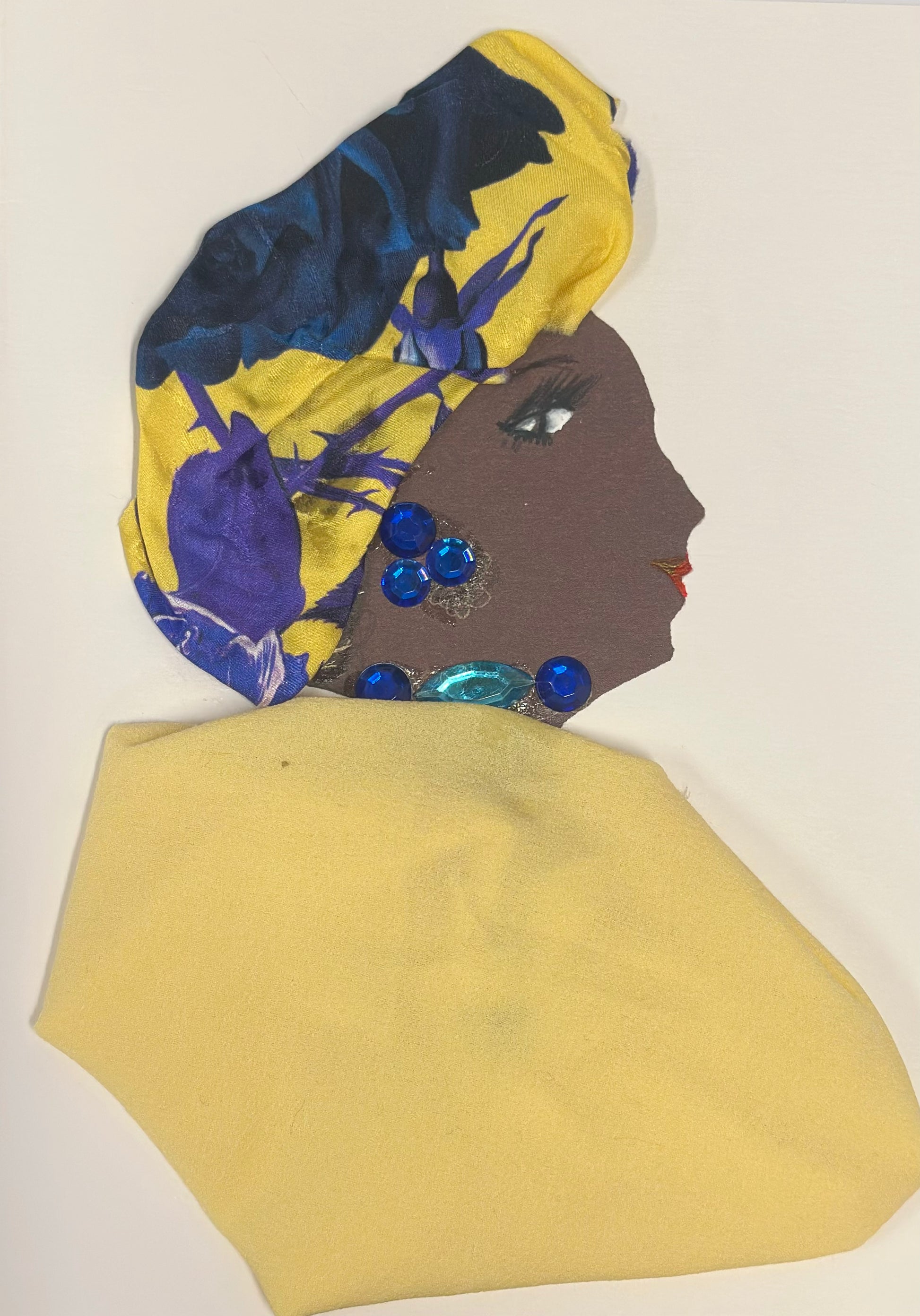 A handmade card displays a side-profile portrait of a woman with deep brown skin, long lashes, and red lips. She wears a yellow headwrap decorated with large blue and purple floral patterns, along with blue gemstone earrings and a coordinating blue gem at her neckline. A soft yellow fabric is draped over her shoulders, completing the vibrant look.
