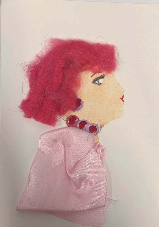 A handmade card shows a side-profile portrait of a woman with soft peach skin and bright, textured pink hair. She wears round red and purple jewel-like earrings and a matching choker, with a pale pink satin fabric draped around her shoulders. Her makeup includes bold eyelashes and red lips, giving her a stylish and expressive appearance.