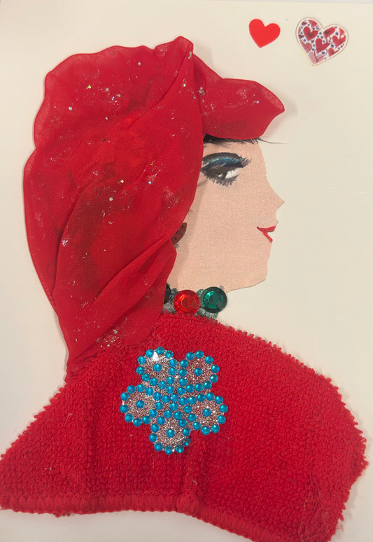 A handmade card features a side-profile portrait of a woman with soft beige skin, dramatic eye makeup, and red lips. She wears a flowing red headscarf and a textured red wrap decorated with a bright blue jeweled flower. Green gem accents sit at her neckline, and two small heart stickers appear above her head.