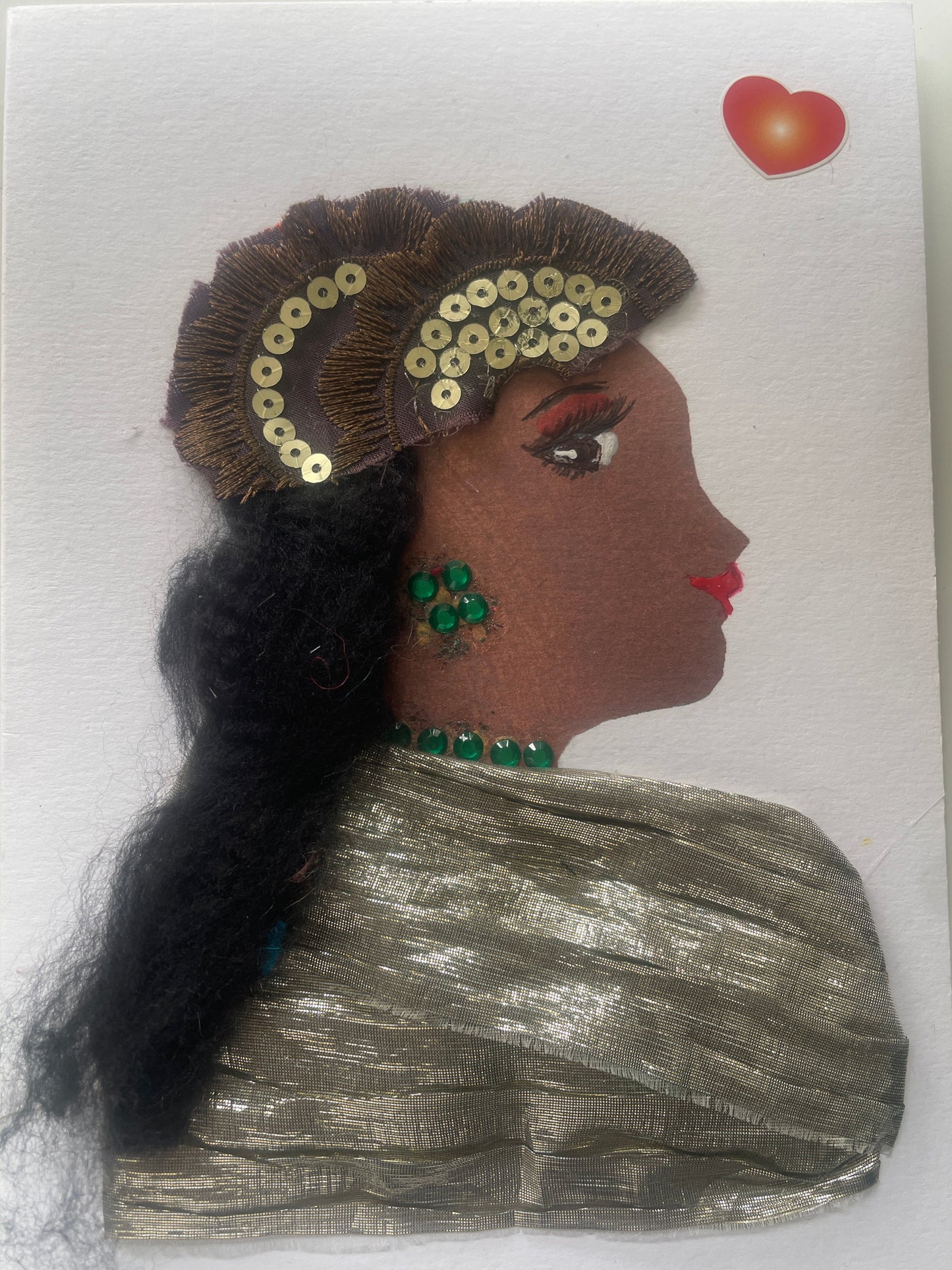 This is a handmade card of a woman named Gilda Goldhawk. She wears a metallic gold blouse, has long black hair with a sequinned headpiece, green gem jewellery, and a red-orange heart sticker.