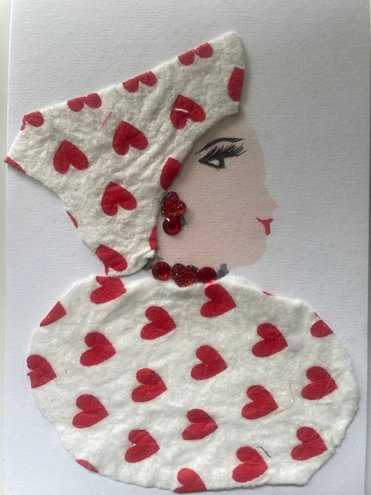 This is a handmade card of a woman named Luma Loxford. She wears a white blouse and wrap with red hearts, red gem jewellery, and a simple background.