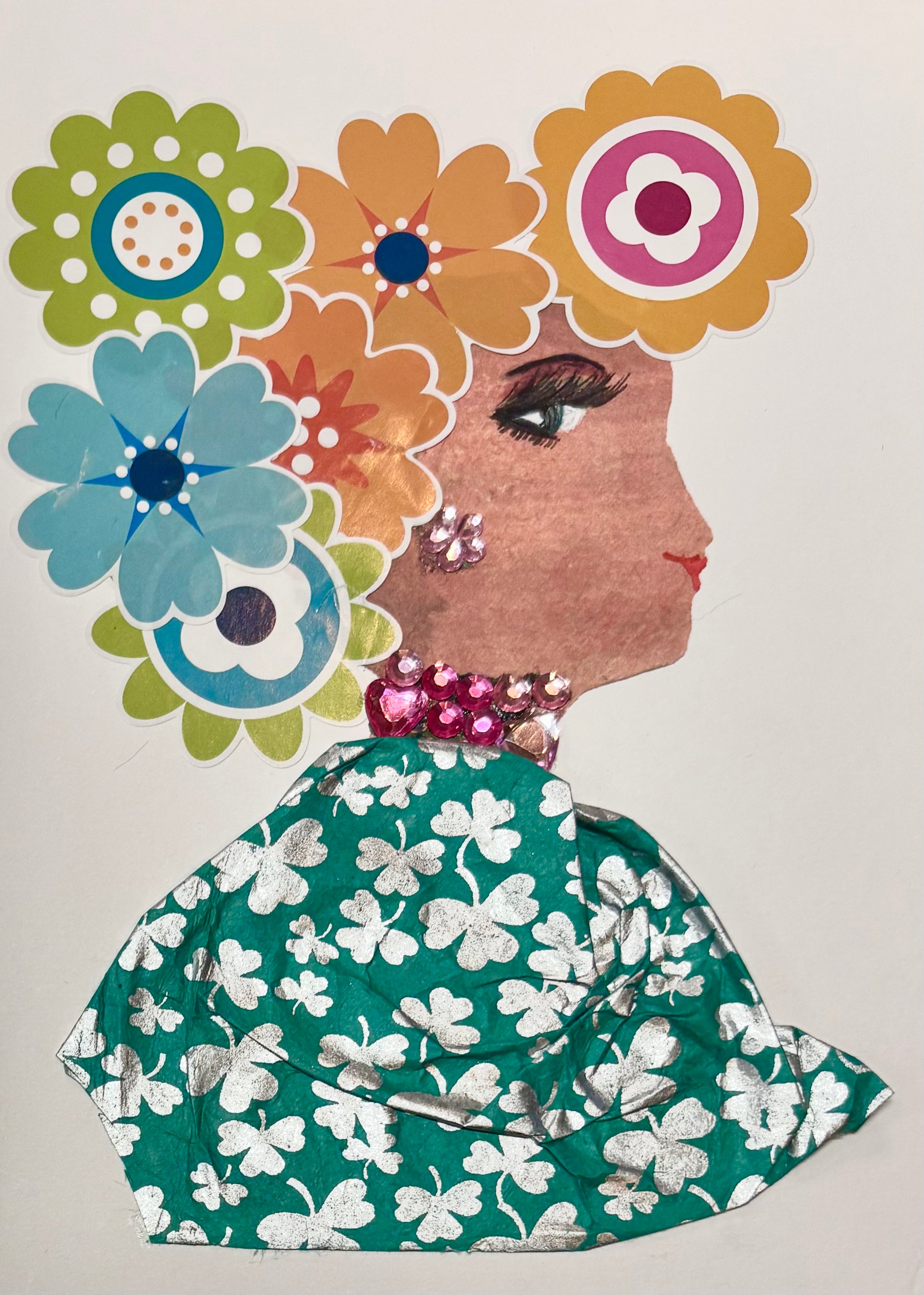 This is a handmade card of a woman named Lady Aldgate. Her hair is made from a colourful arrangement of large paper flowers in orange, blue, green, yellow, and pink. She wears sparkly pink gem earrings and a matching necklace. Her outfit is created from green fabric with a bold silver clover pattern.