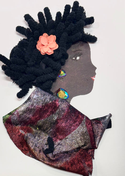 This is a handmade card of a woman named Kira Kensington. Her hair is made from thick black yarn arranged in curls, with a pink flower on one side. She has two shiny, colourful earrings. A soft, shiny fabric in red and purple tones is wrapped around her shoulders like a scarf. Her face is drawn in profile with a calm expression.